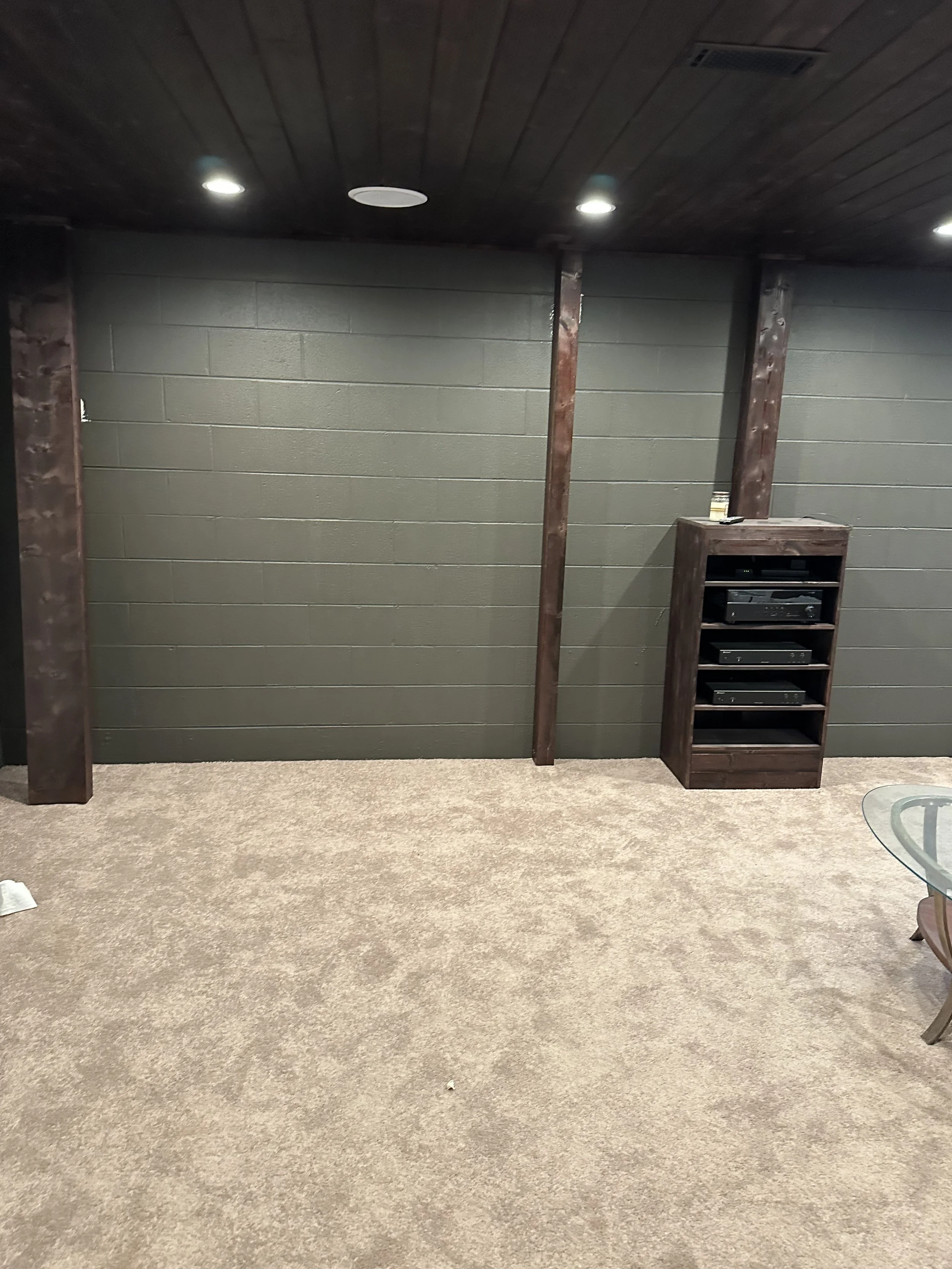 An empty room with a beige carpet, gray brick wall, and ceiling with recessed lighting. There is a small wooden rack or cabinet on the right side of the wall. Part of a glass table is visible in the bottom right corner.