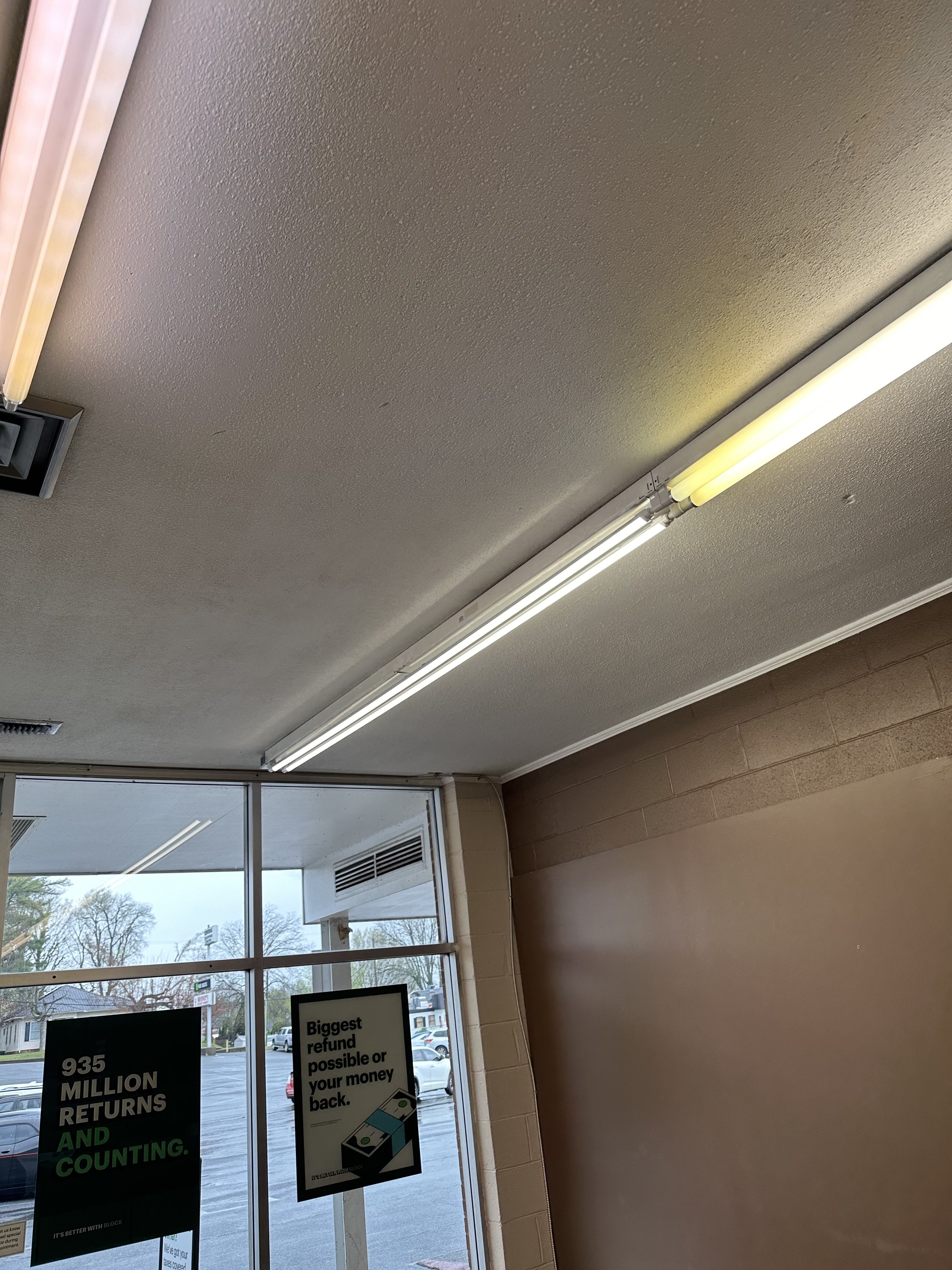 Indoor view of a building's interior showing part of the ceiling with fluorescent lights, a window, and some signs on the wall near the cash register area.