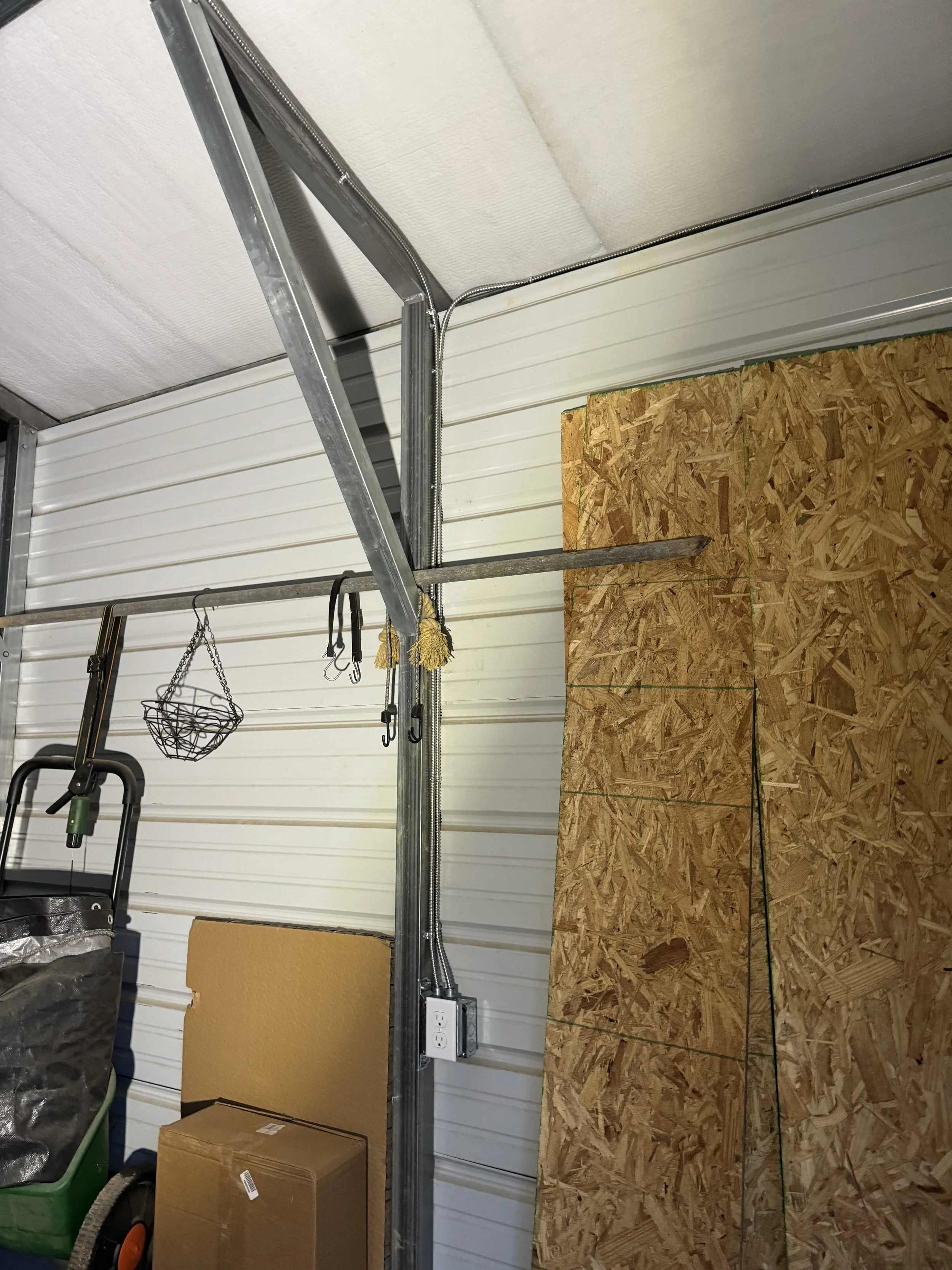 Interior view of a garage or workshop with OSB plywood on the floor, a metal track or framing, coiled electrical conduit, hanging hooks, a power strip, a cardboard box, and various garage items.
