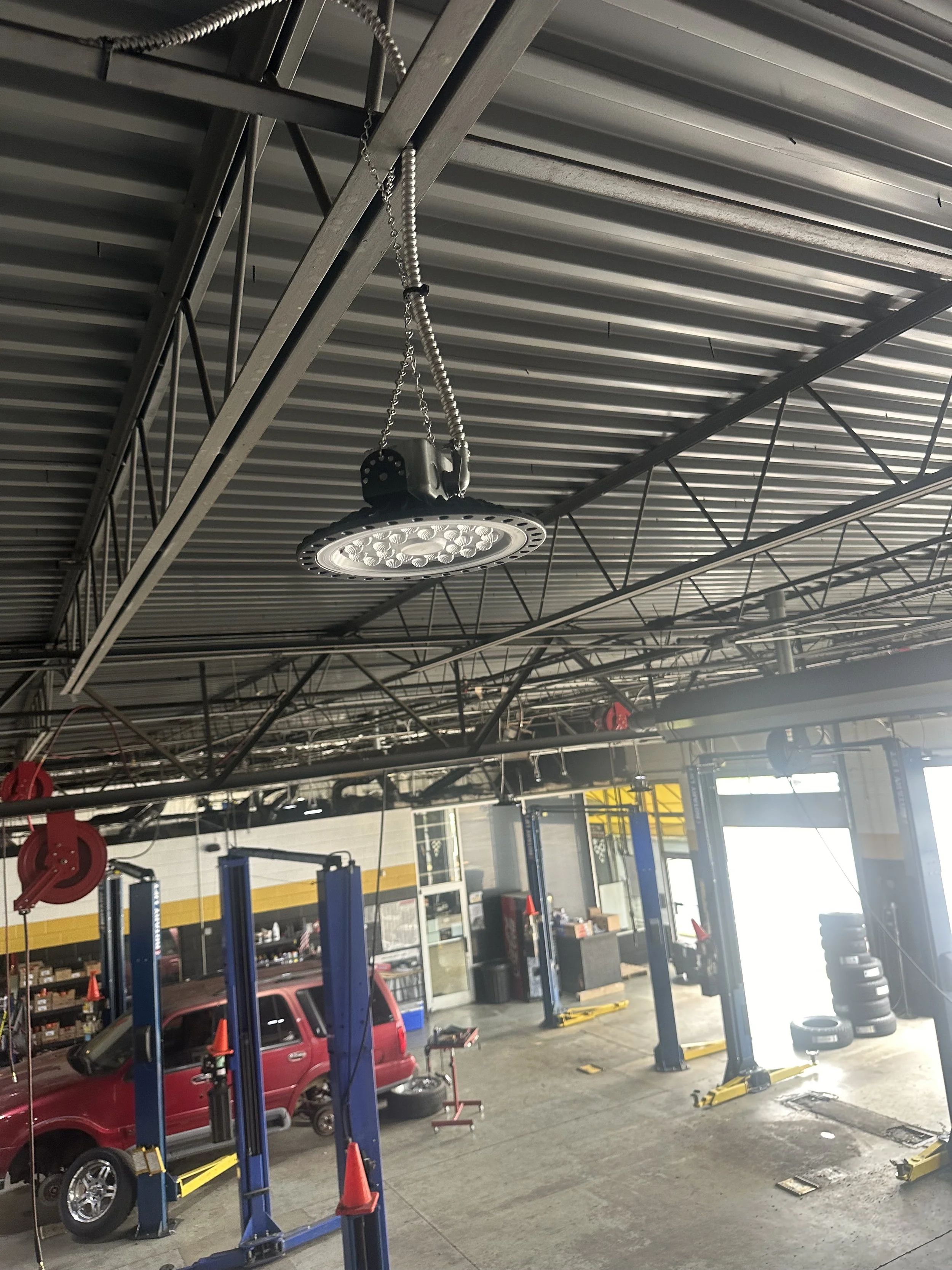 Inside an automotive repair shop with a red vehicle on a lift, tire stacks, and various tools and equipment in the background. The ceiling has industrial lighting and metal support beams.