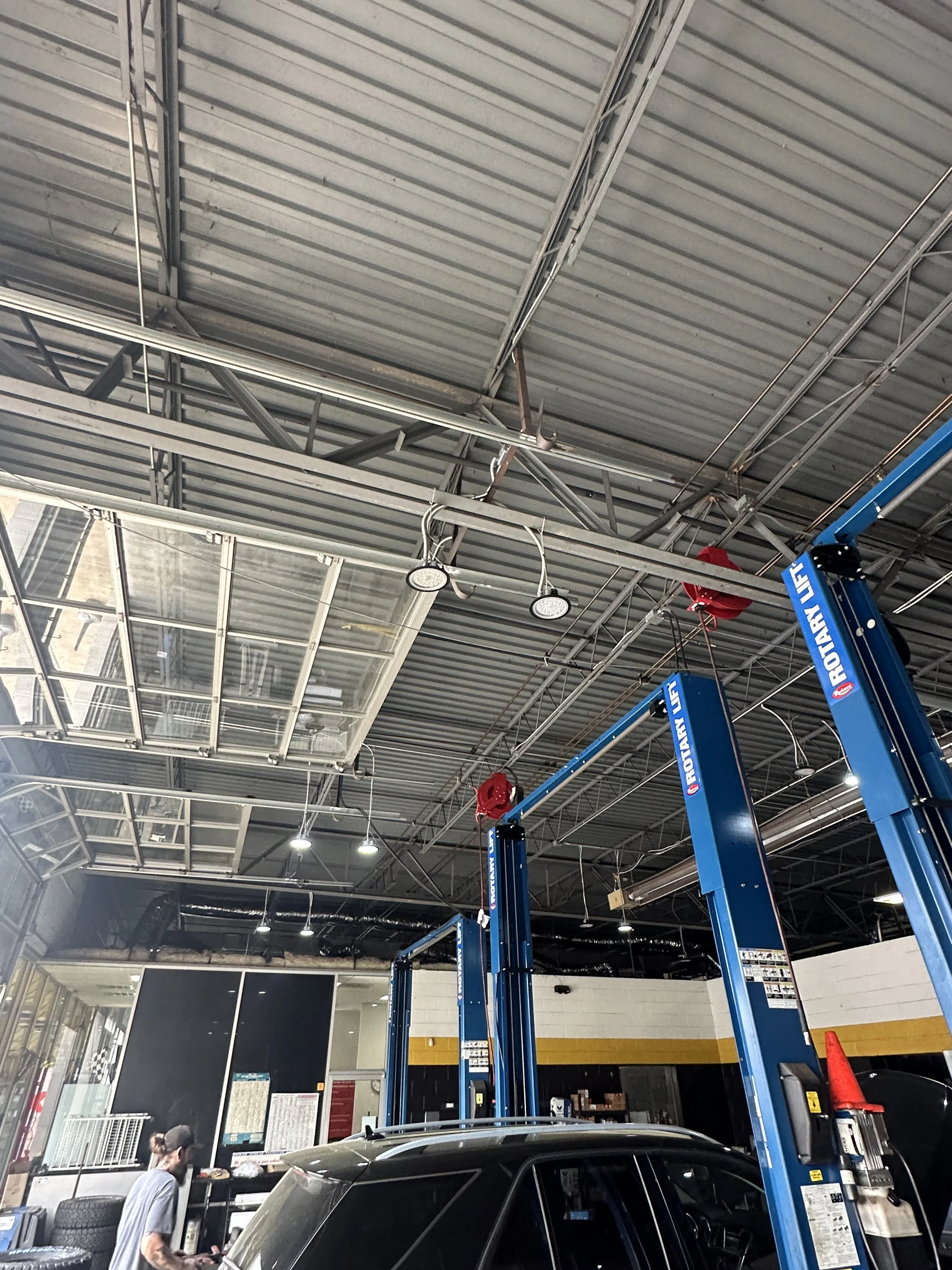 Inside a car repair shop, blue automotive lifts, a black car, and a person wearing gray shirt and black pants near tools and tires.