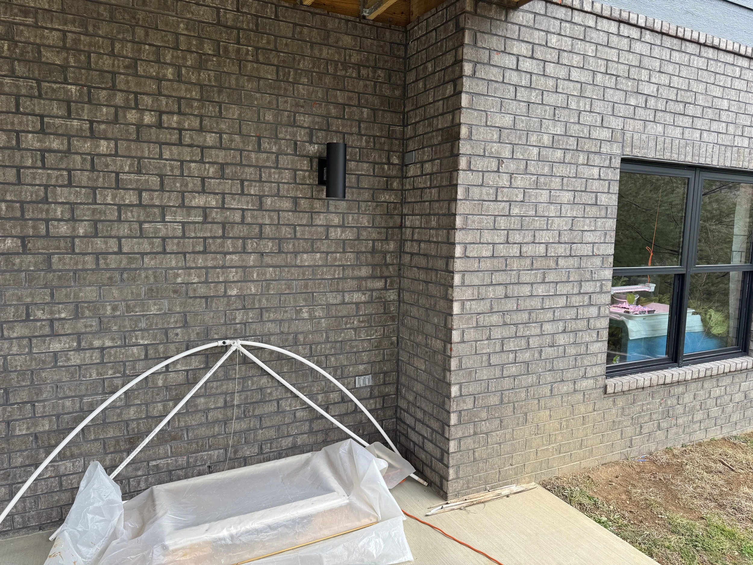 Exterior of a house under construction with brick walls and a window, construction materials and tools, including a plastic sheet and a white pipe, are visible on the ground.