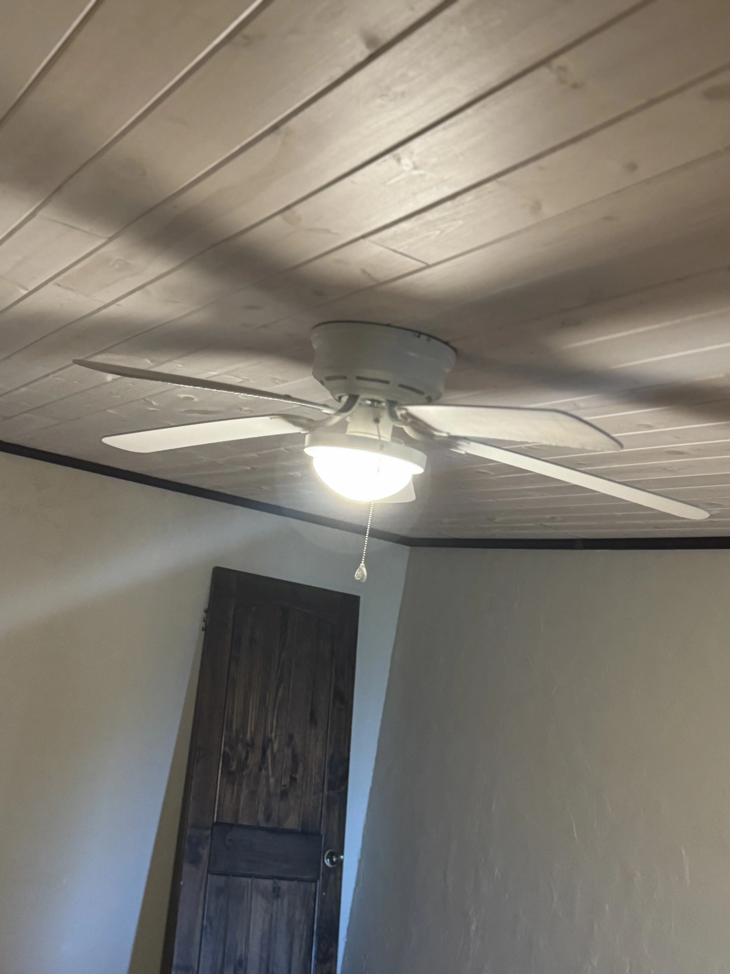 Ceiling fan and light fixture with pull chain, mounted on wooden ceiling, in a room with light gray walls and a dark wooden door.