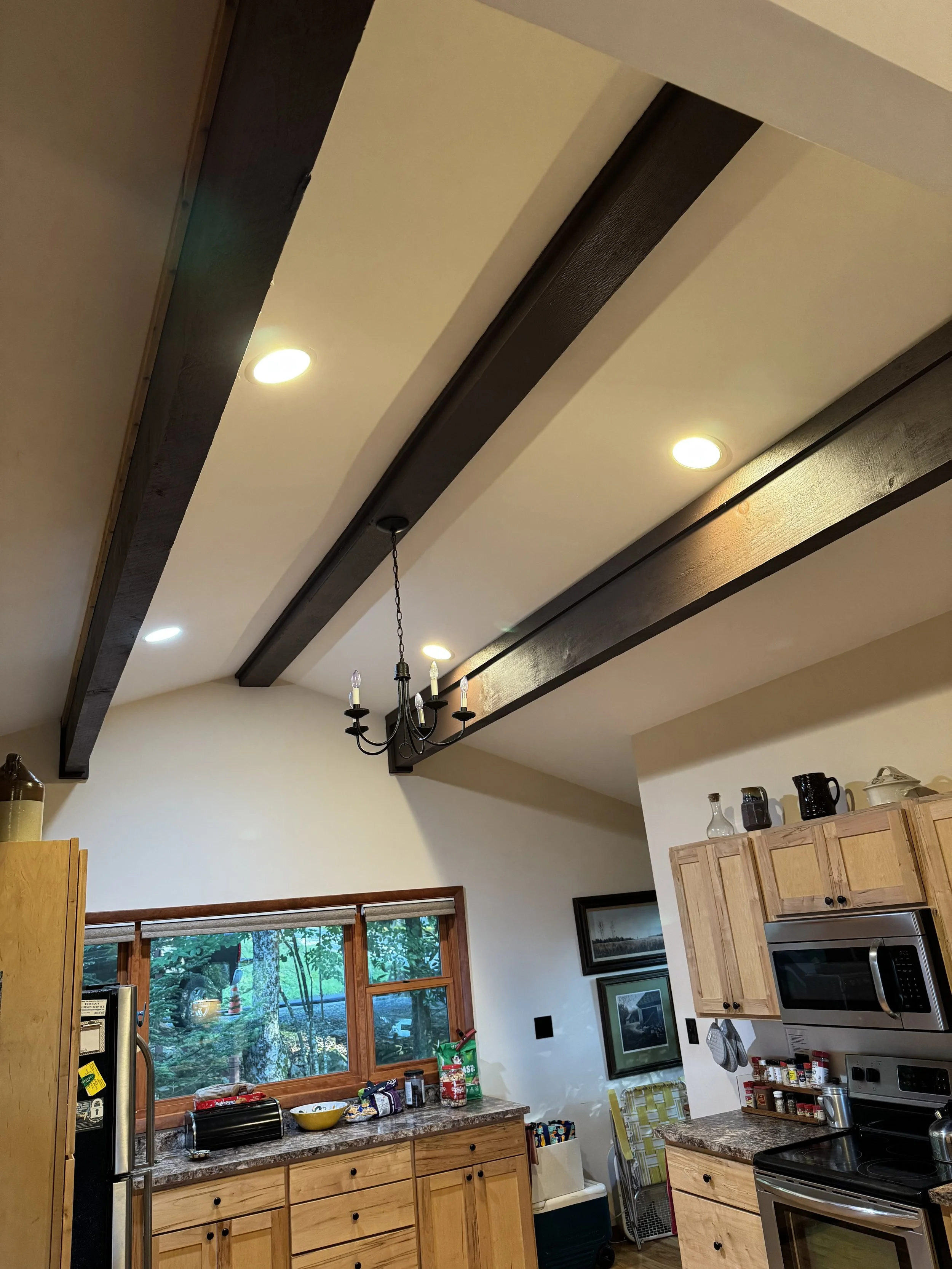 Kitchen ceiling with wooden beams, recessed lighting, and a chandelier, viewed from below. Kitchen cabinets, a window with a view outside, and various kitchen appliances and items are visible.