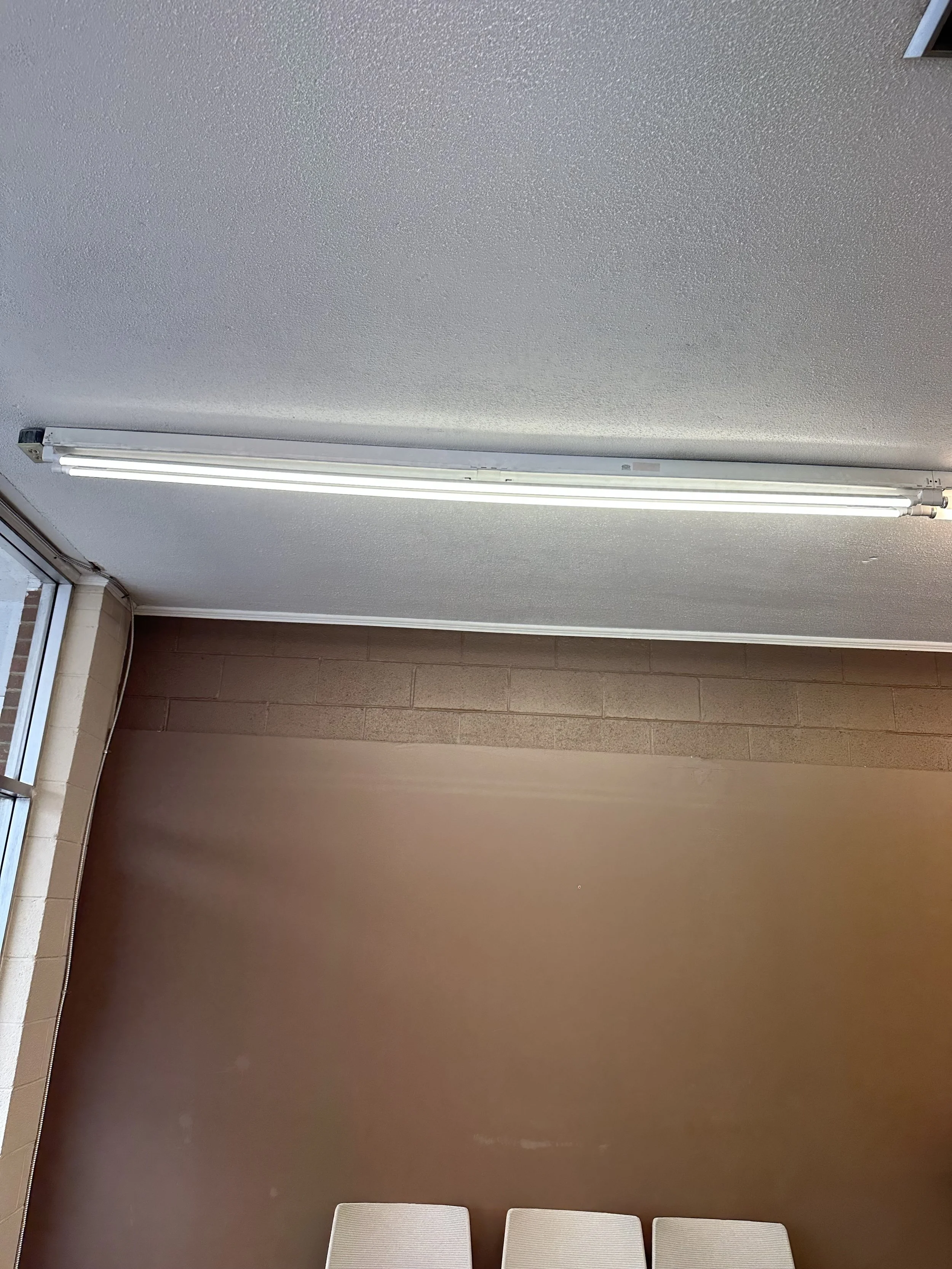 Interior ceiling with fluorescent lights, brown wall, beige chairs, and part of a window.