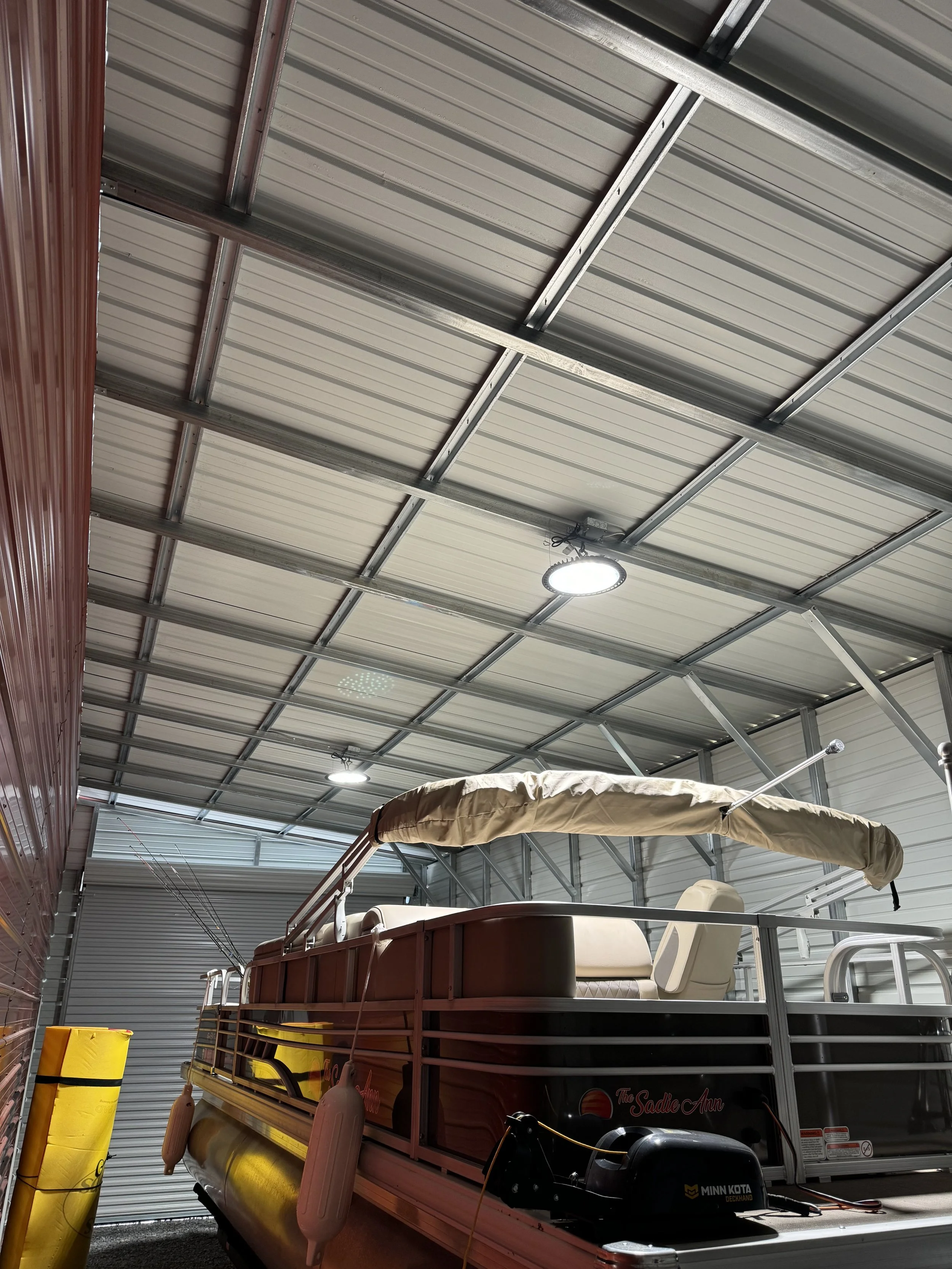 A boat with beige seating and a beige canopy stored inside a metal building with a corrugated metal roof, fluorescent lighting, and garage door.