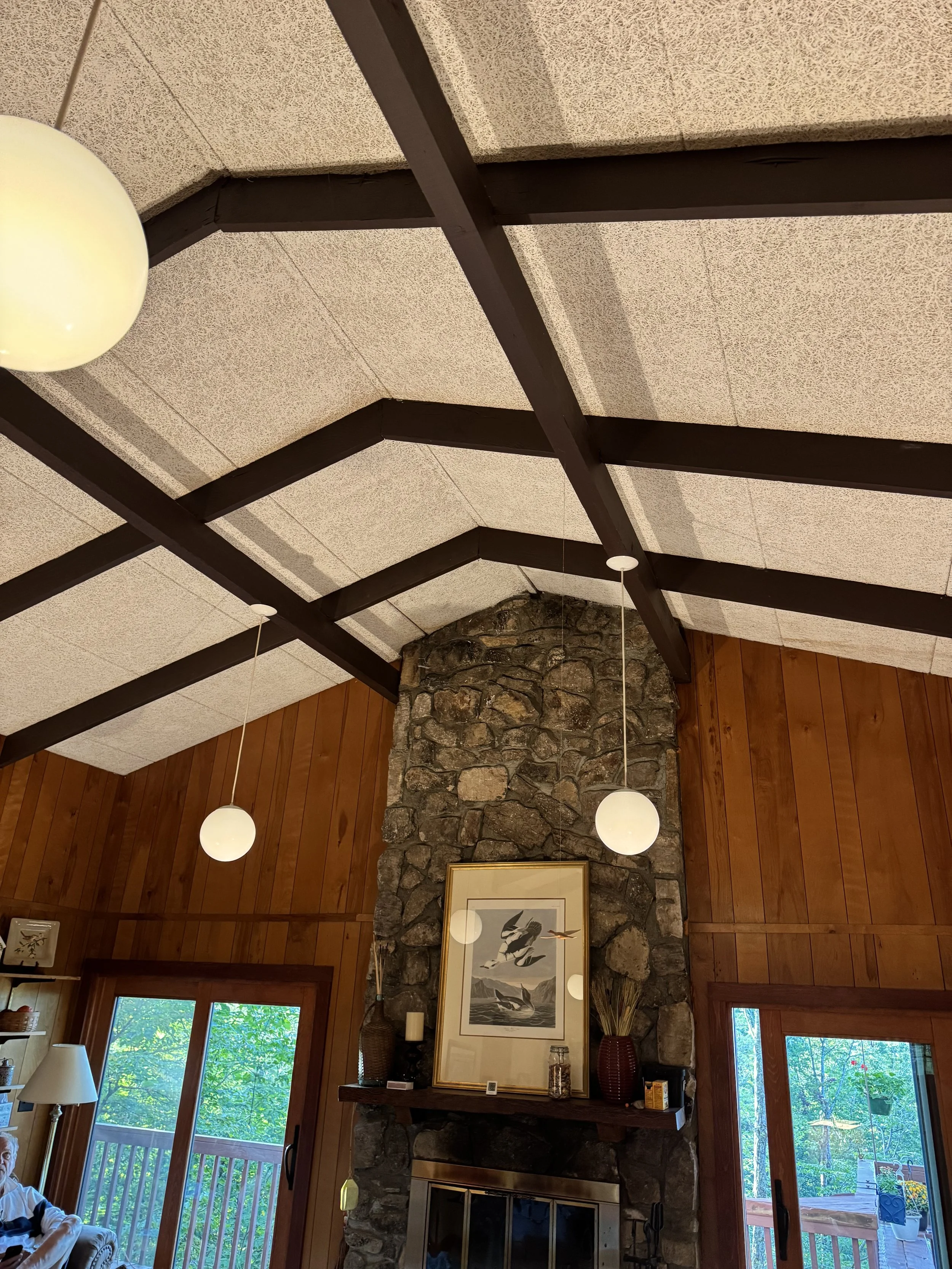 Living room with a stone fireplace, wooden walls, and large windows showing trees outside. Decor includes framed artwork above the fireplace, vases, and a chandelier with globe lights hanging from the ceiling.