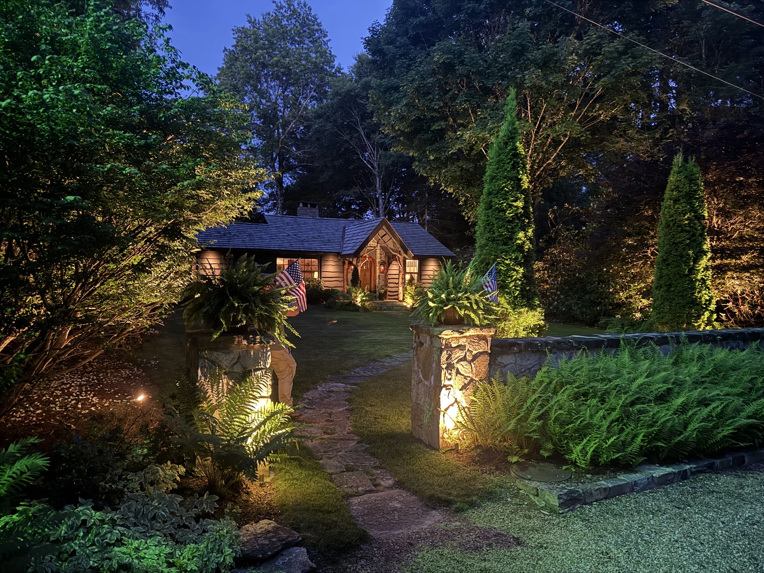 Nighttime view of a garden with a small house illuminated from within, surrounded by lush green trees and plants, with outdoor lighting accenting pathways and a stone pillar.