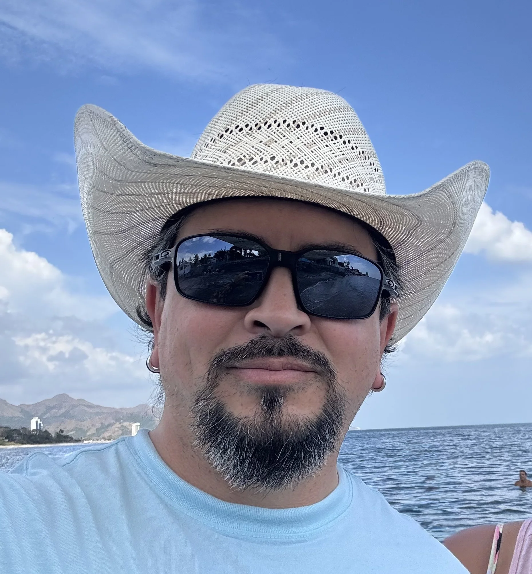 A person wearing a wide-brimmed white straw cowboy hat and black sunglasses, near the water and mountains, with a blue sky and some clouds in the background.