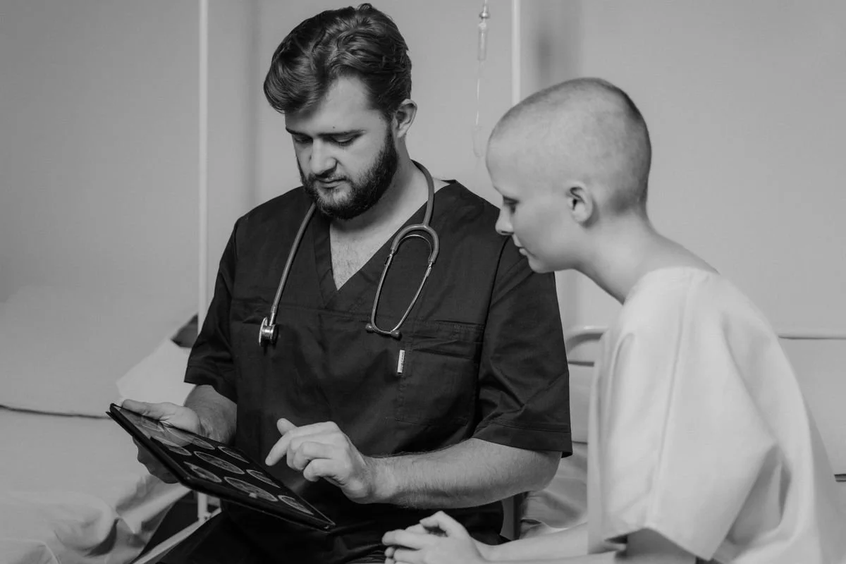 A male healthcare professional in scrubs looking at a tablet screen with a young patient who has a shaved head, in a hospital room.
