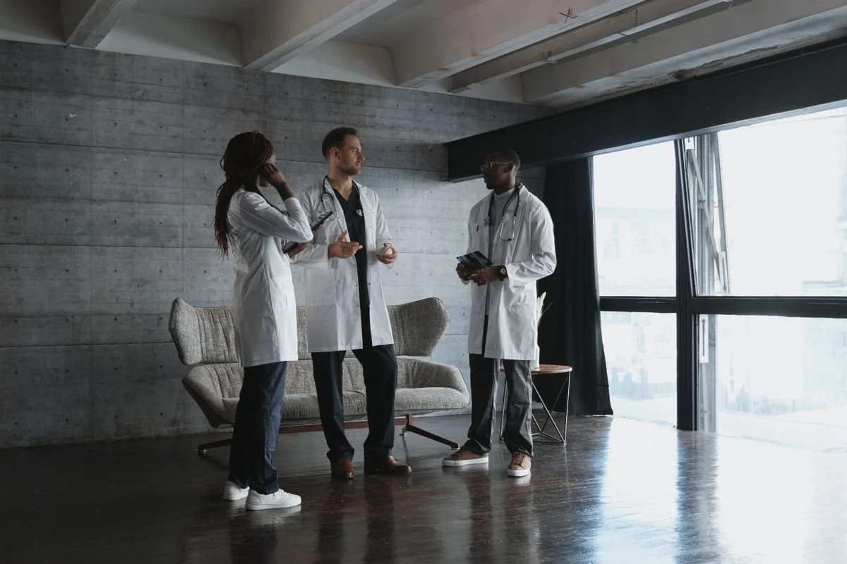 Three healthcare professionals, two men and one woman, standing and talking in a modern hospital or clinic lounge area with a concrete wall and large window.