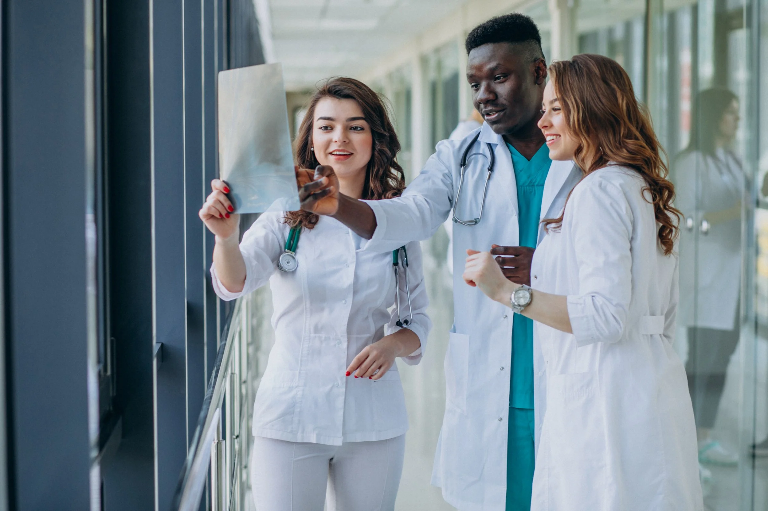 Group of three healthcare professionals, two women and one man, looking at an X-ray inside a modern hospital corridor with large windows.