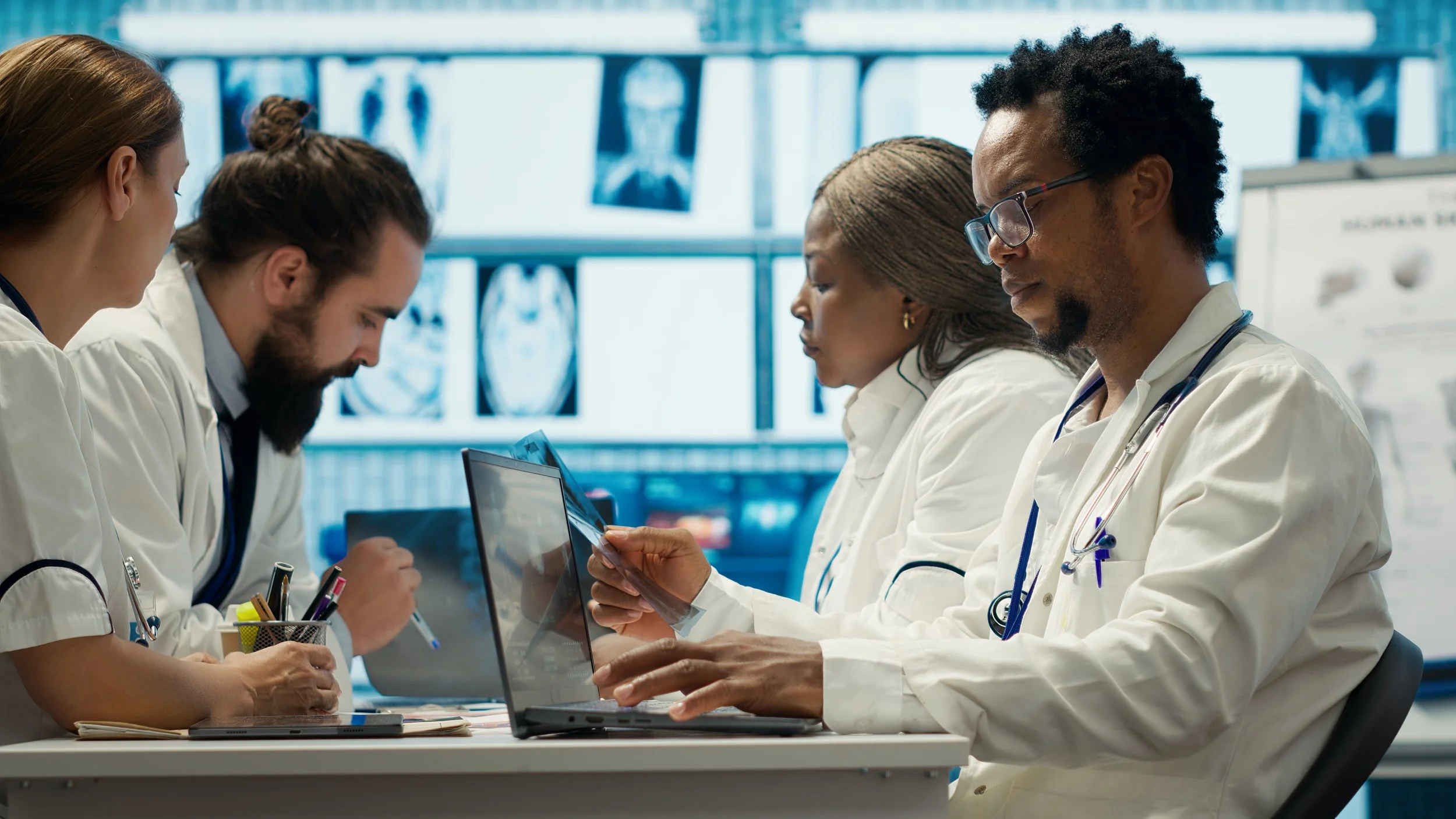 Group of medical professionals, including women and men of diverse backgrounds, sitting at a table with laptops, medical charts, and writing utensils, engaged in discussion in a hospital or clinic setting with medical images displayed on screens in the background.