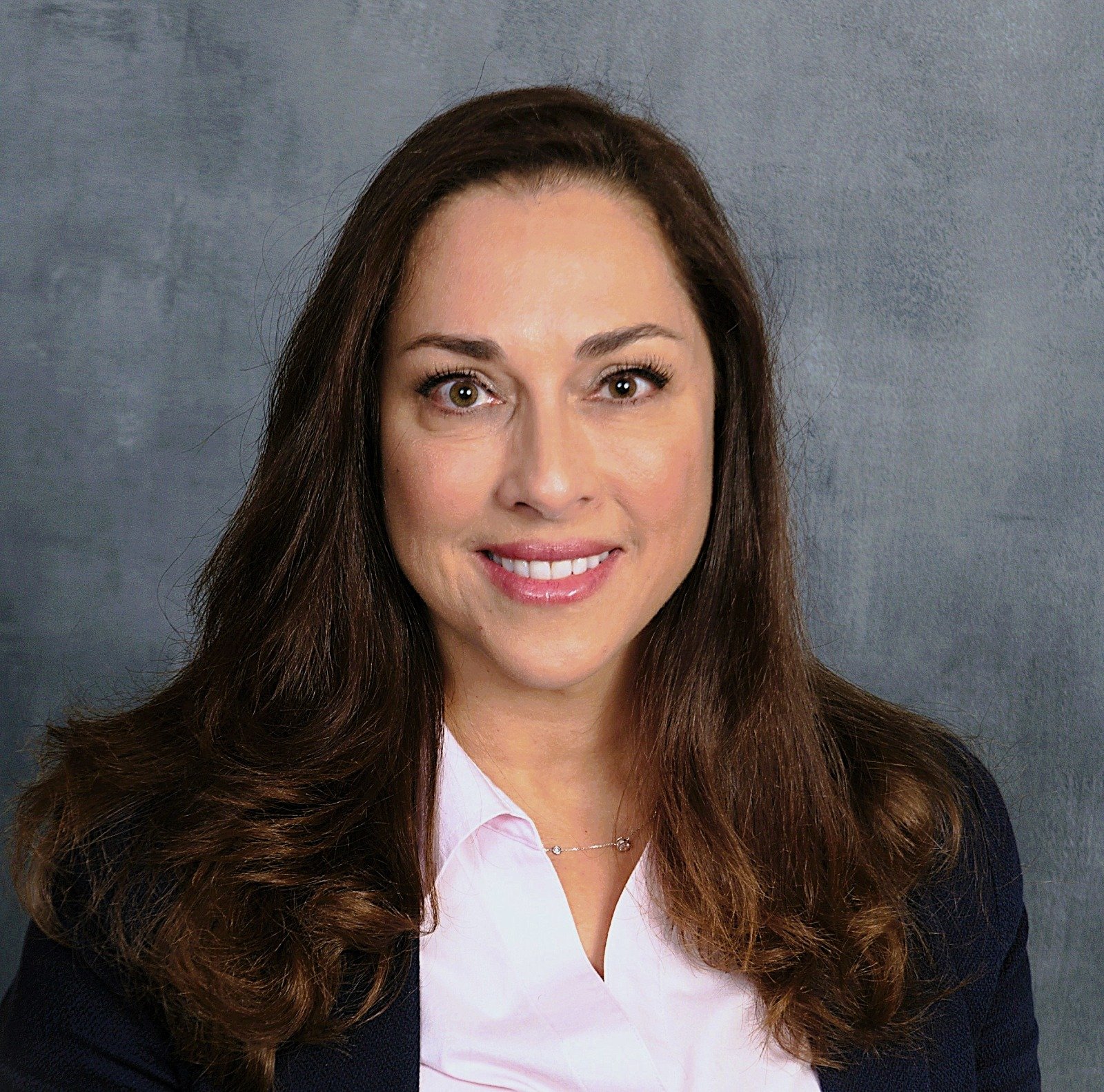A woman with long, wavy brown hair smiling and looking at the camera, wearing a white shirt and dark blazer against a grey background.