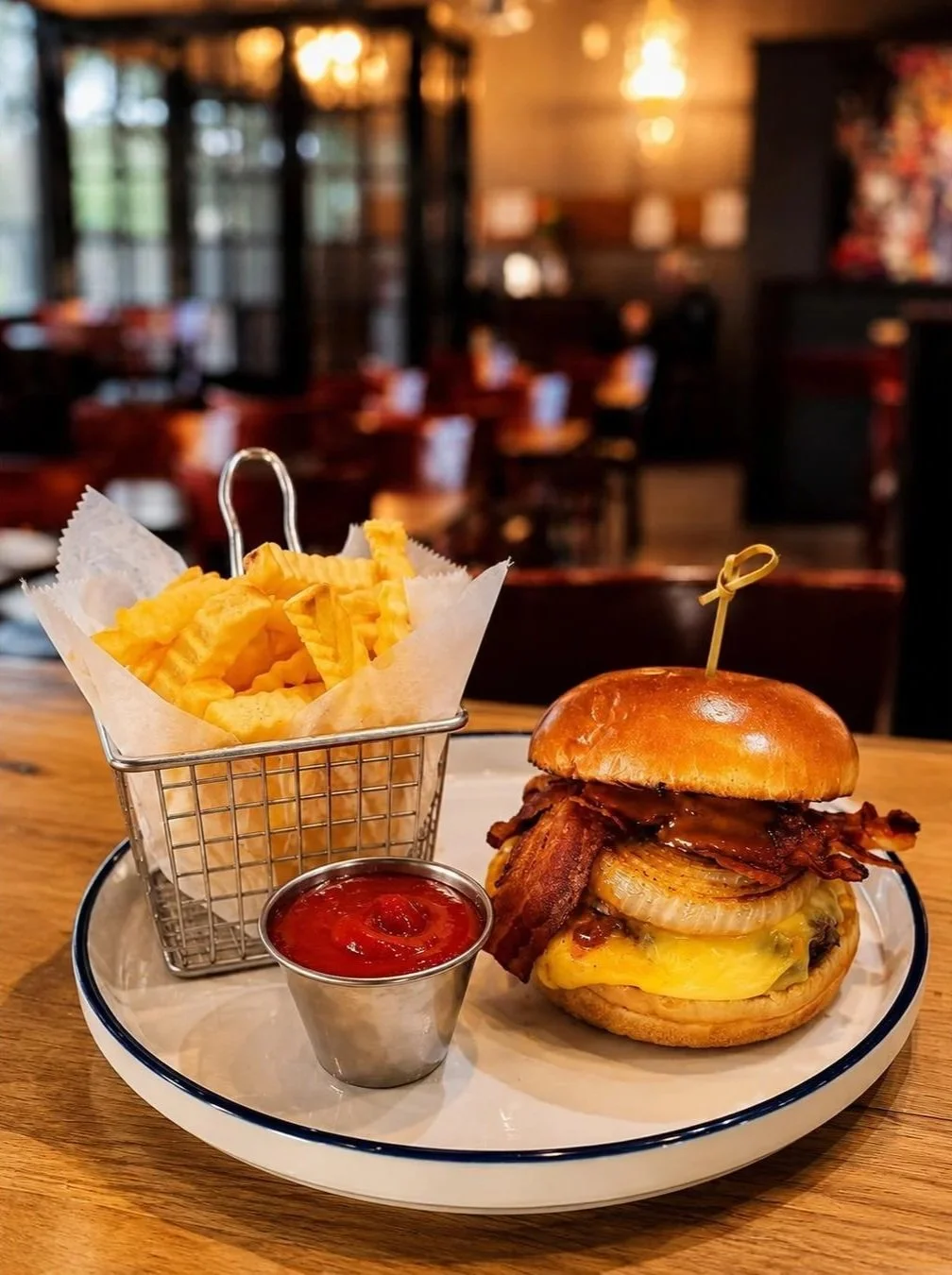 A cheeseburger with bacon, grilled onion, and melted cheese on a bun, served with fries and ketchup in a small metal cup, on a white plate in a restaurant setting.