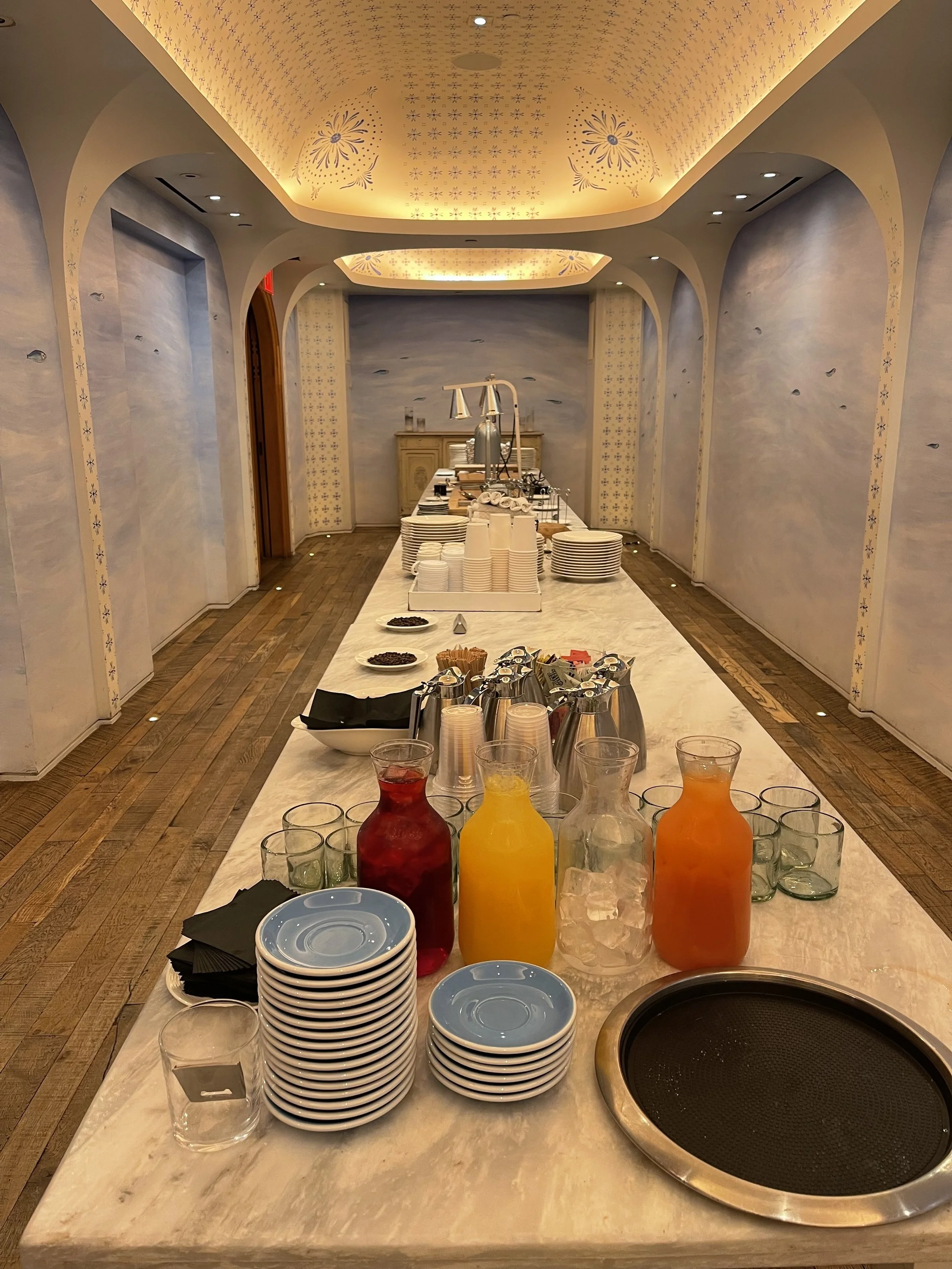 A buffet setup with stacked plates, glasses, pitchers of colored drinks, and containers of food and condiments on a marble table. There is a hot plate at the front and a background with a decorative curved ceiling with lights and fish-themed wall acc