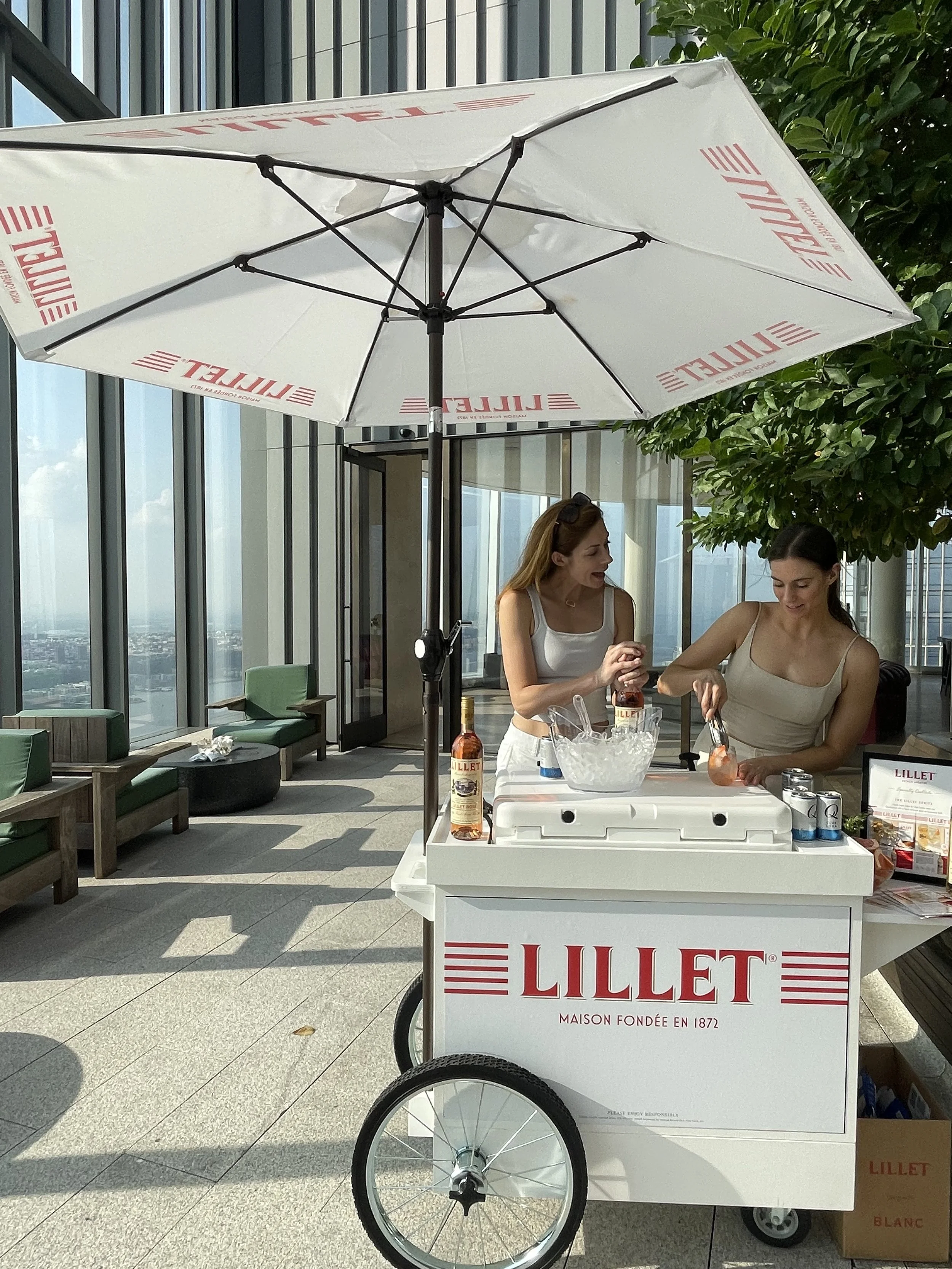 A white Lilyet ice cream cart with two large wheels, topped with a white umbrella, set on an outdoor building terrace with green chairs and a view of the city.