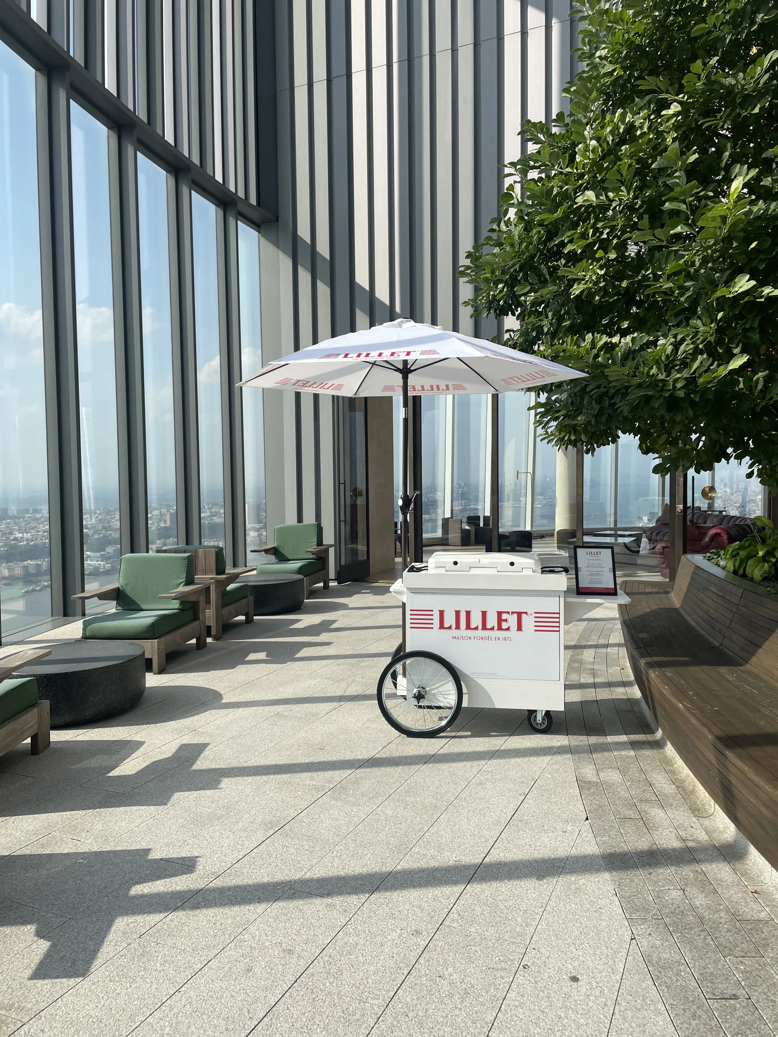 Modern rooftop indoor lounge with large windows, green cushioned seats, a green leafy plant, and a Lillet-branded portable cart with an umbrella.
