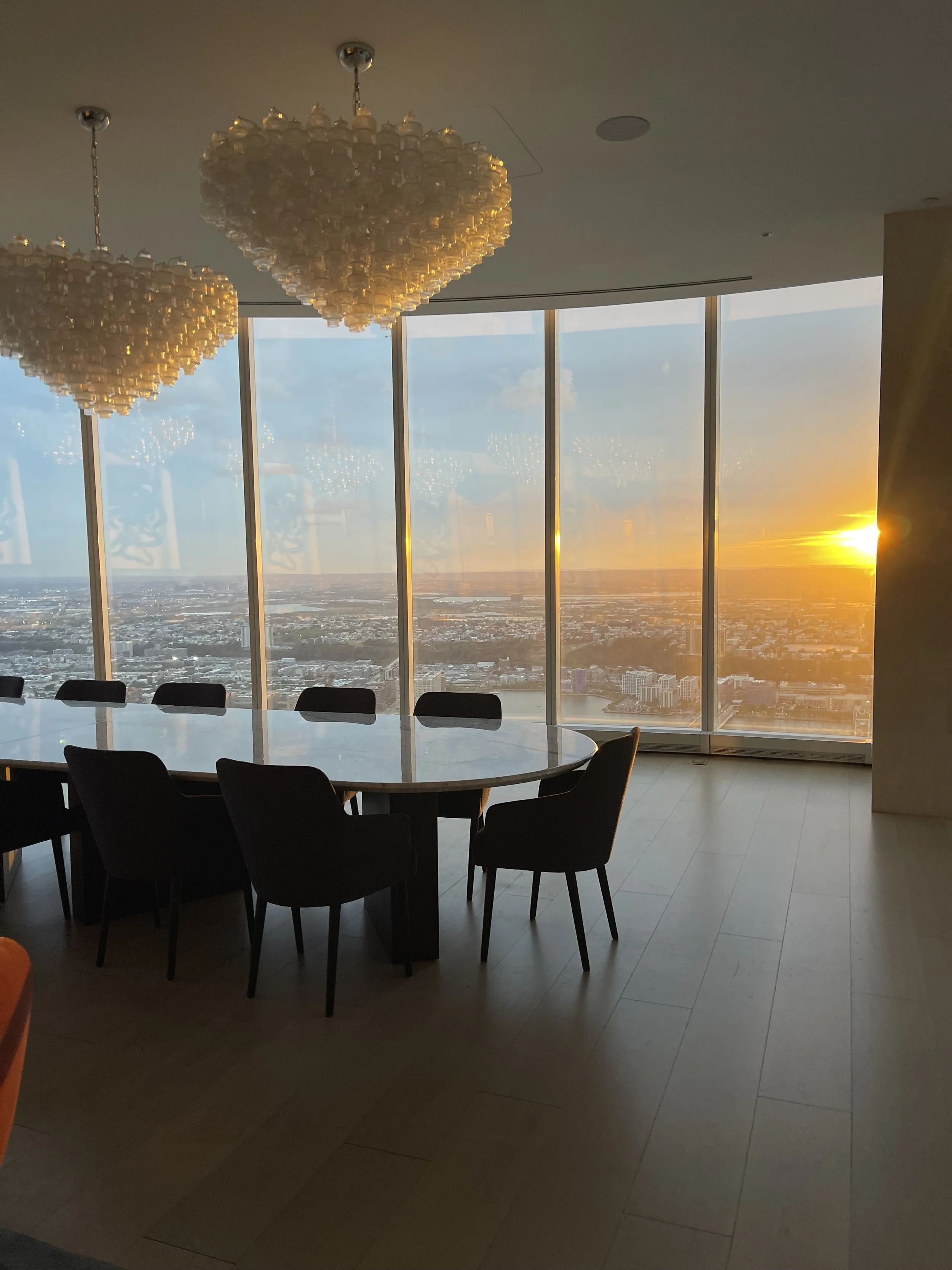Interior of a high-rise building with a large round table and chairs, panoramic windows showing a cityscape at sunset, two chandeliers hanging from the ceiling.