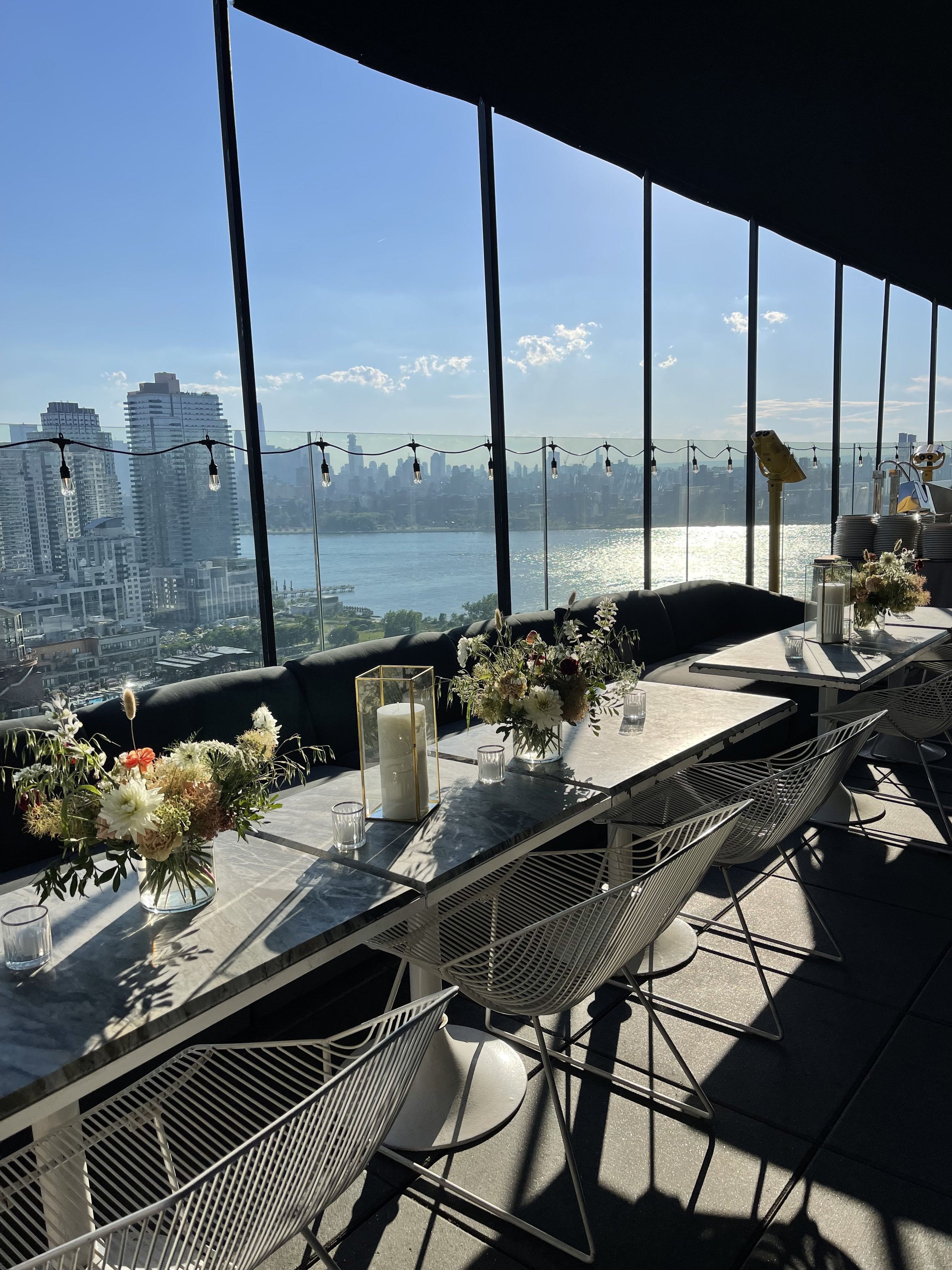 A sunny indoor space with a view of a city skyline and water through large glass windows, decorated with flower arrangements and candles on a marble table.