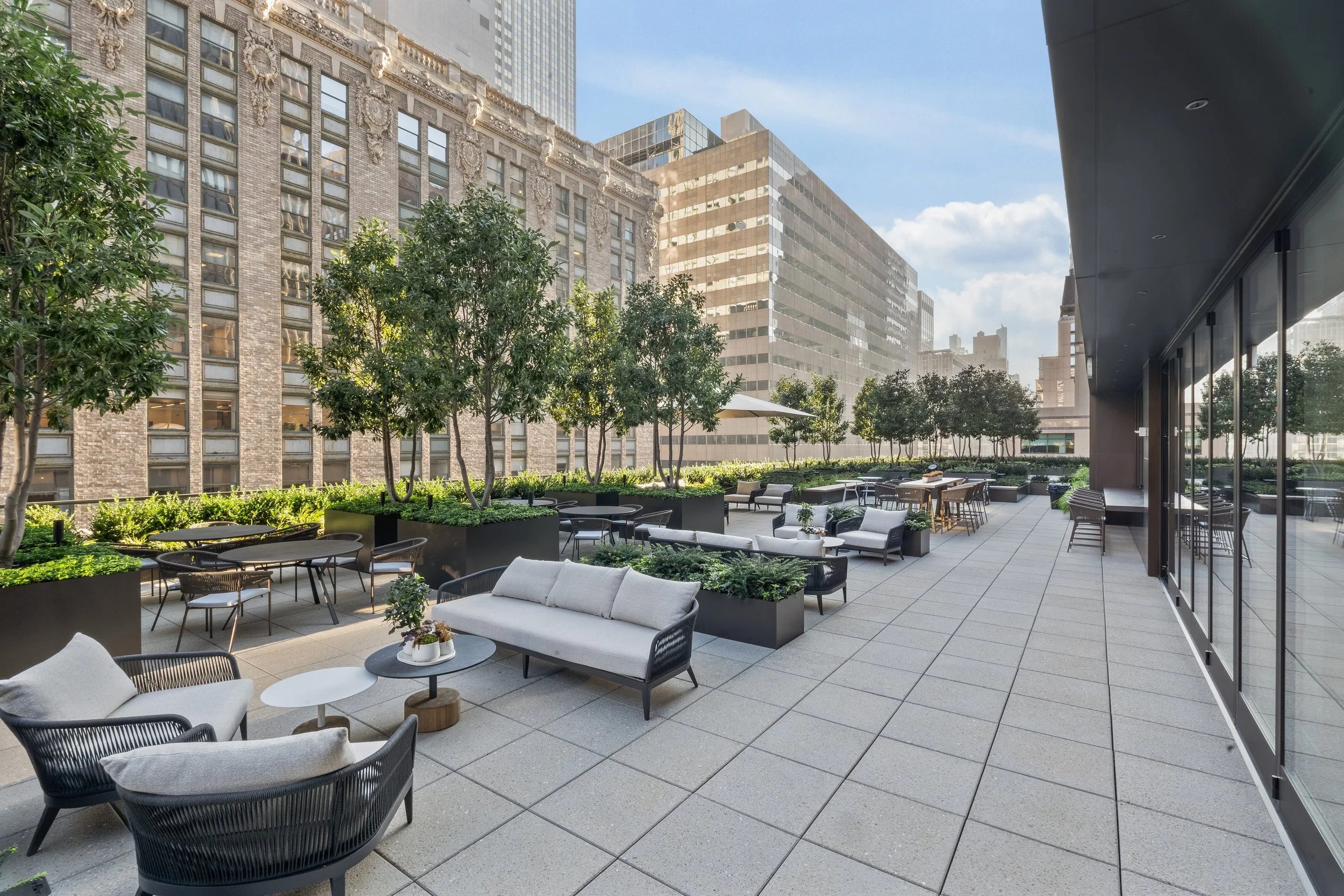 An outdoor terrace with modern furniture and potted plants, surrounded by tall buildings in an urban cityscape.