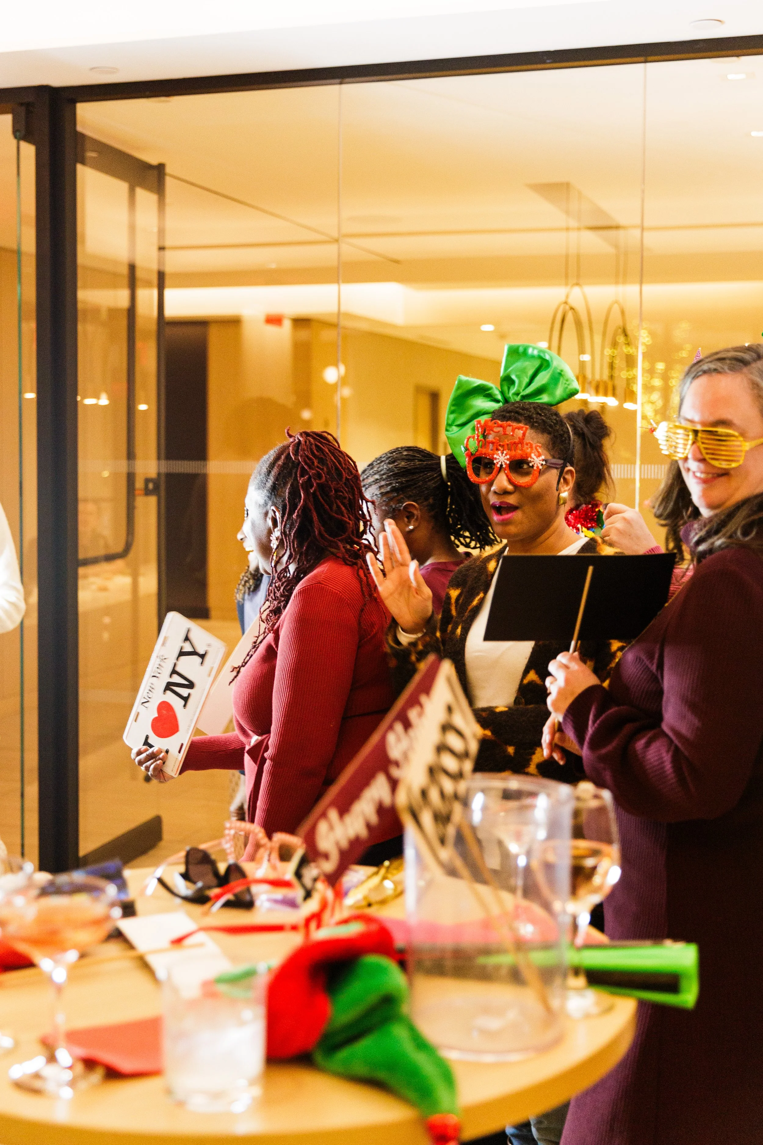Group of people wearing festive accessories at a holiday gathering, standing near a table with Christmas decorations and signs.