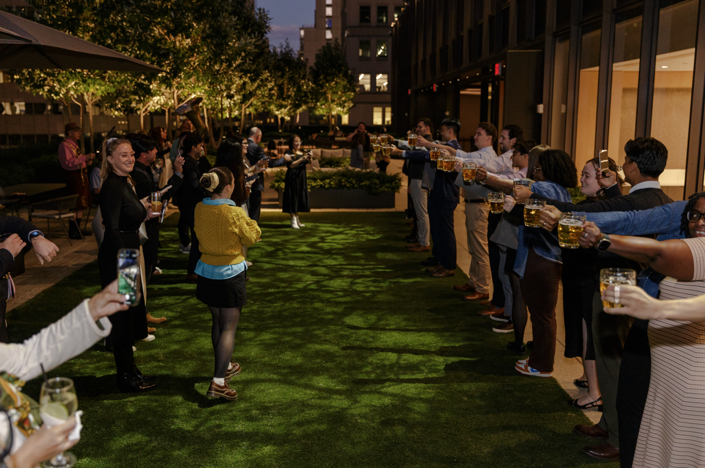 People standing in two lines on an outdoor terrace at night, raising glasses of beer for a toast, with trees and city buildings in the background.
