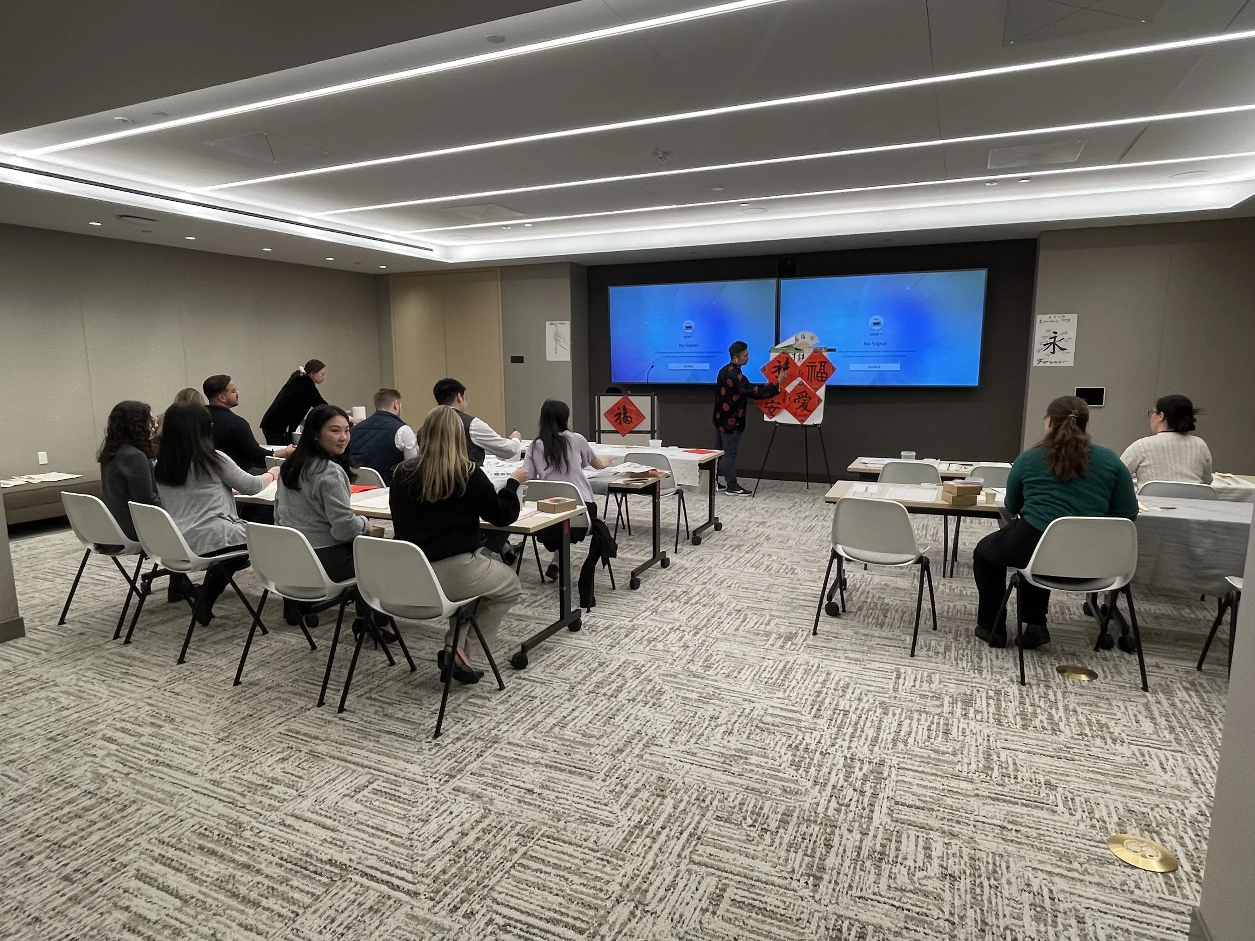 People sitting around tables in a conference room, facing a presenter standing next to a large digital screen displaying a blue background with no signal message, during a presentation or workshop.