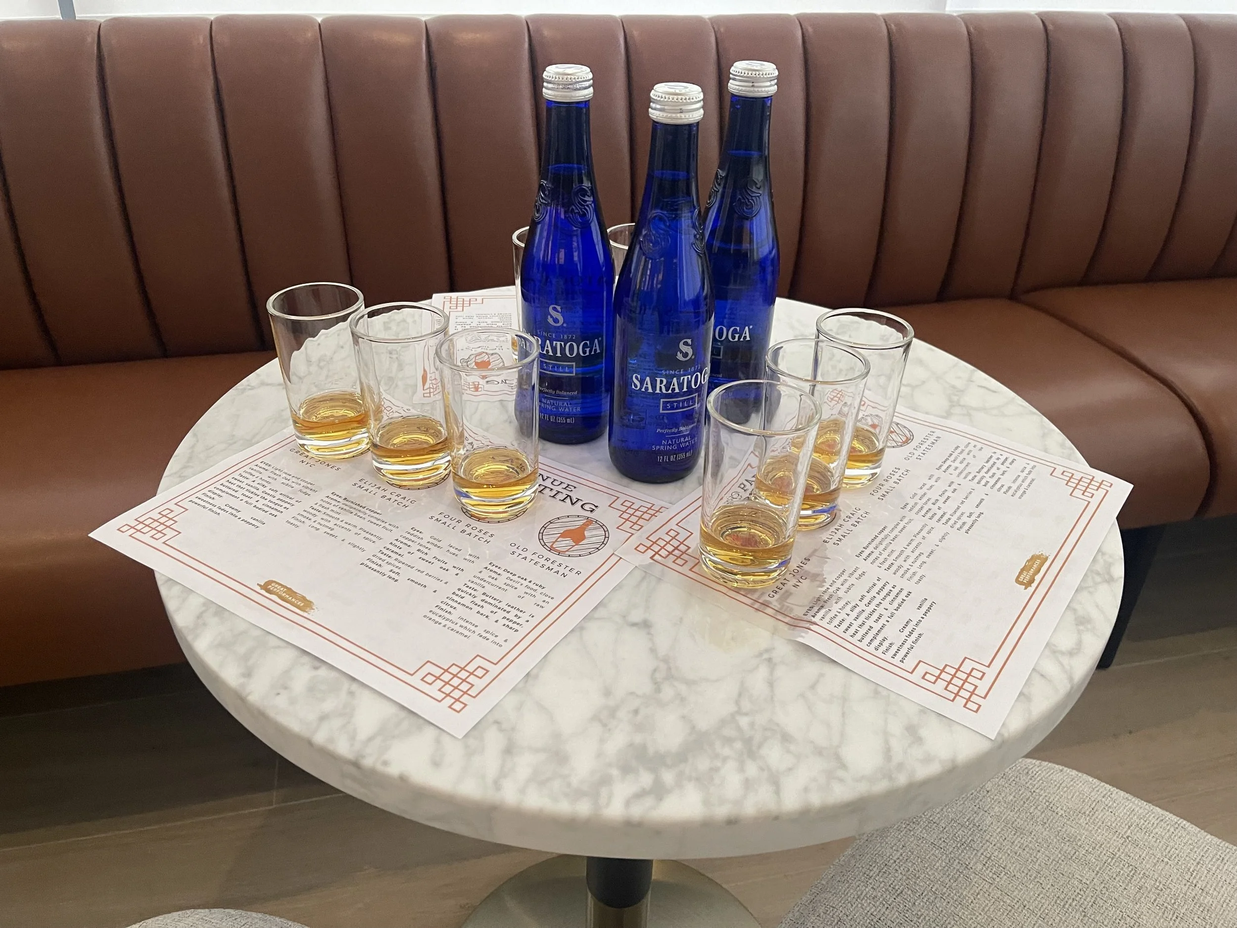 A round white marble table with four glasses filled with amber-colored liquid, three blue glass water bottles labeled Saratoga, and printed menus or placemats, against a brown leather padded bench seat.