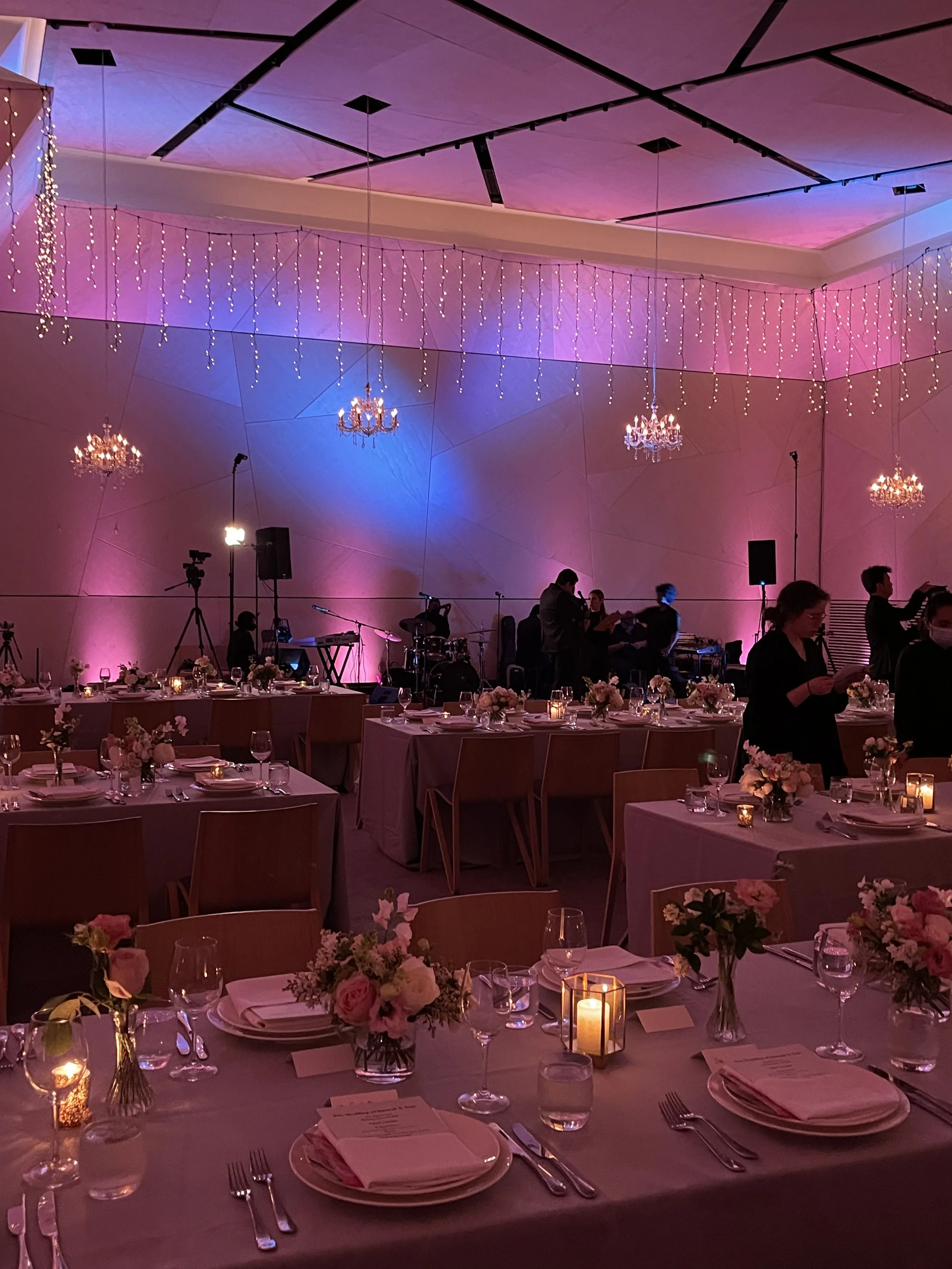 A beautifully decorated banquet hall with tables set for a formal event, featuring floral centerpieces, candles, and elegant tableware. The room has pink and purple lighting, chandeliers hanging from the ceiling, and a stage with musical instruments 