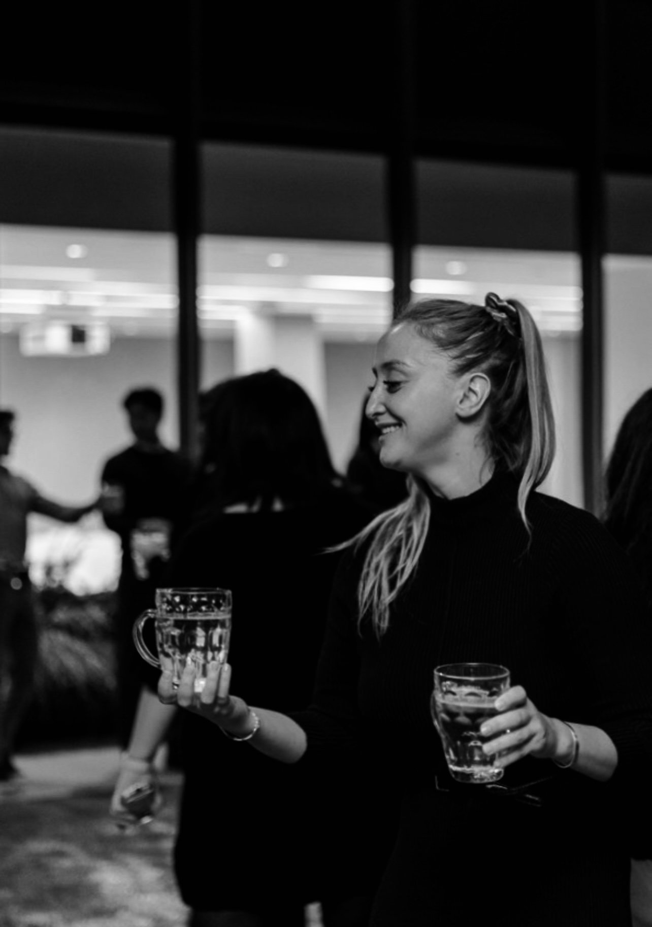 A young woman with long blonde hair tied back in a ponytail, wearing a black top, smiling, and holding two beer glasses at a social gathering in an indoor setting.