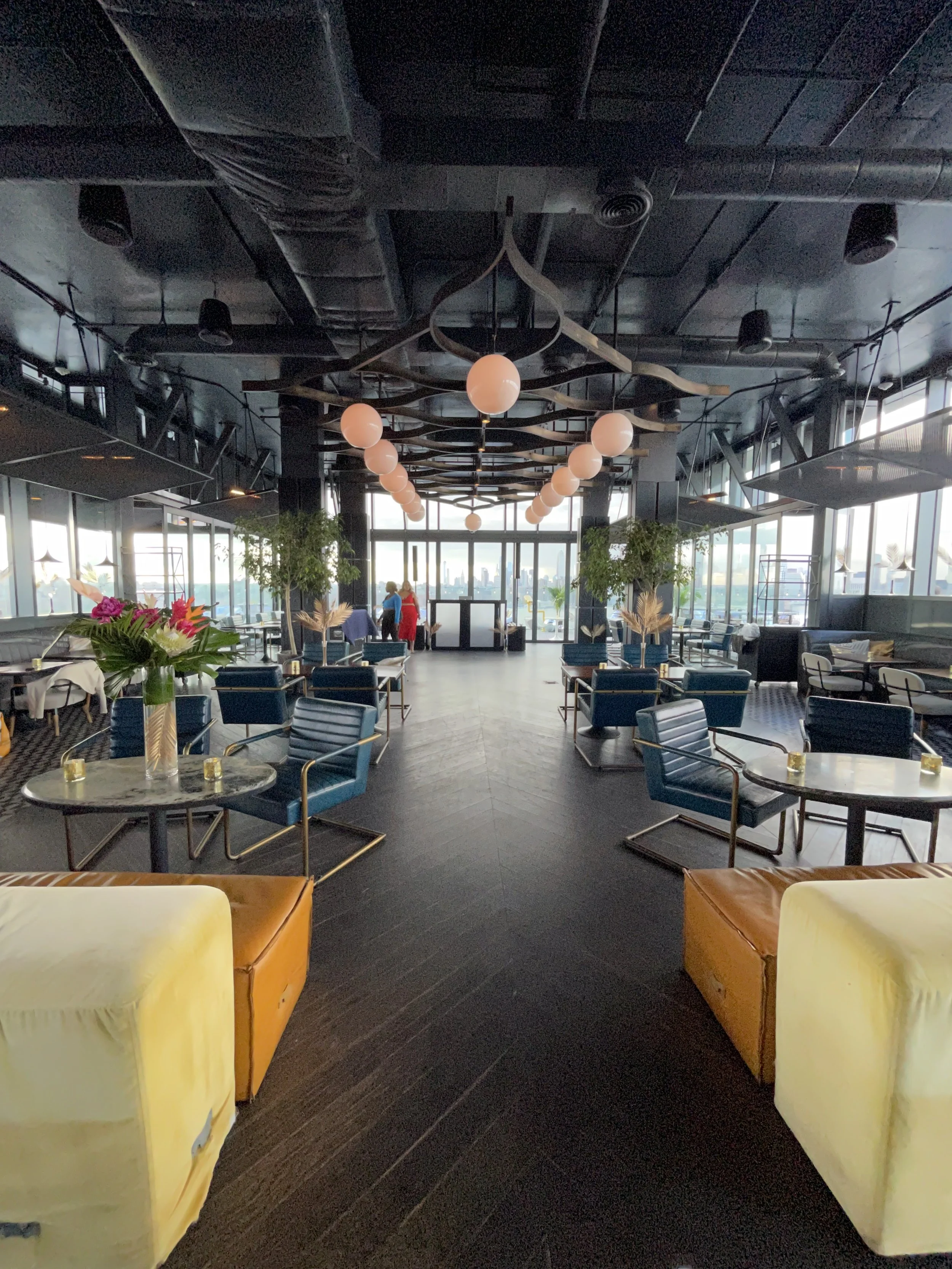 Interior of a modern restaurant or lounge with dark ceiling, wrap-around windows, navy blue chairs, and yellow armchairs, decorated with flower arrangements and pendant lighting.