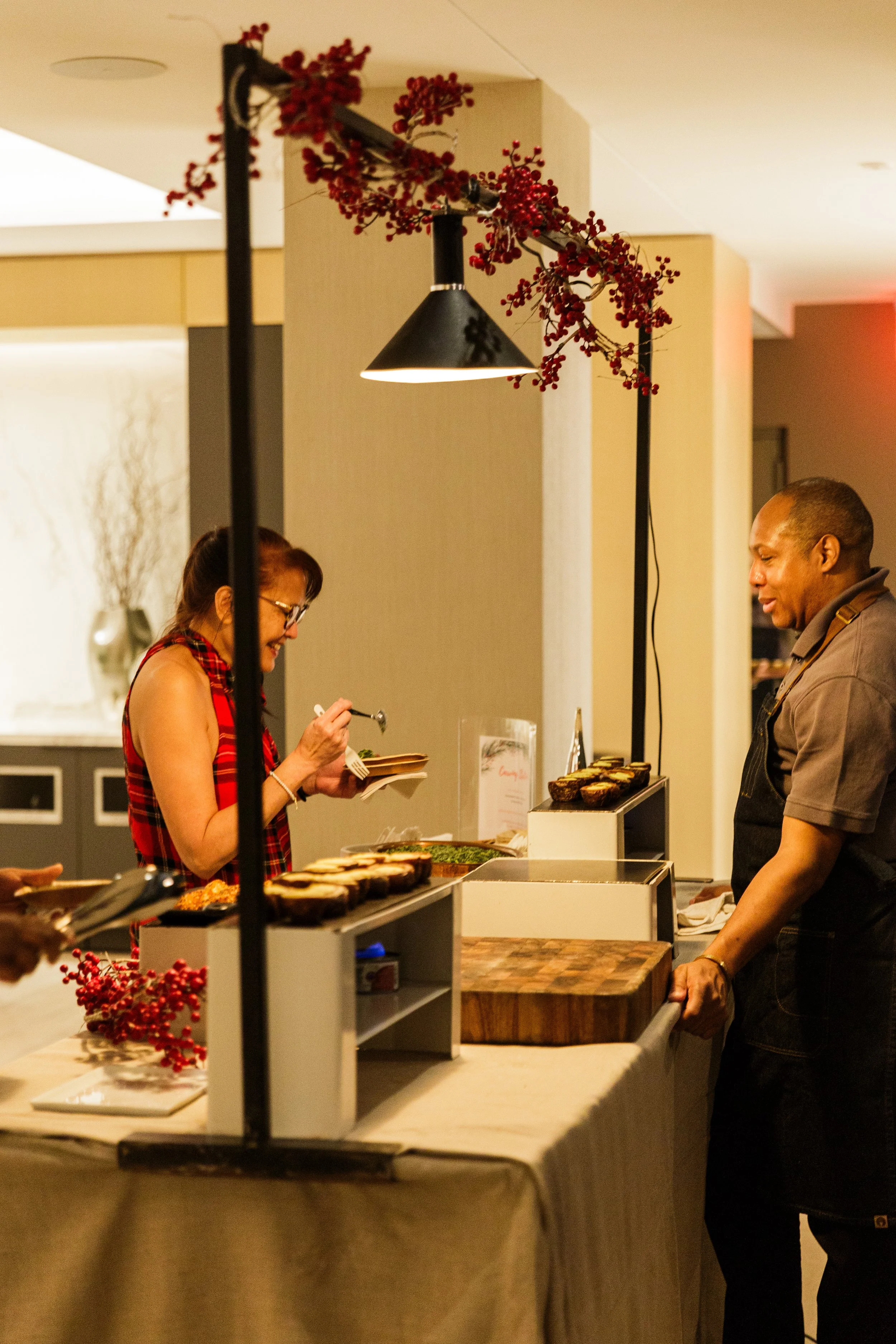A woman with glasses in a red plaid dress, holding a plate and serving herself food, is at a buffet counter. A man in a brown shirt is on the other side of the counter, preparing to serve himself. The buffet table has various dishes including sliced 