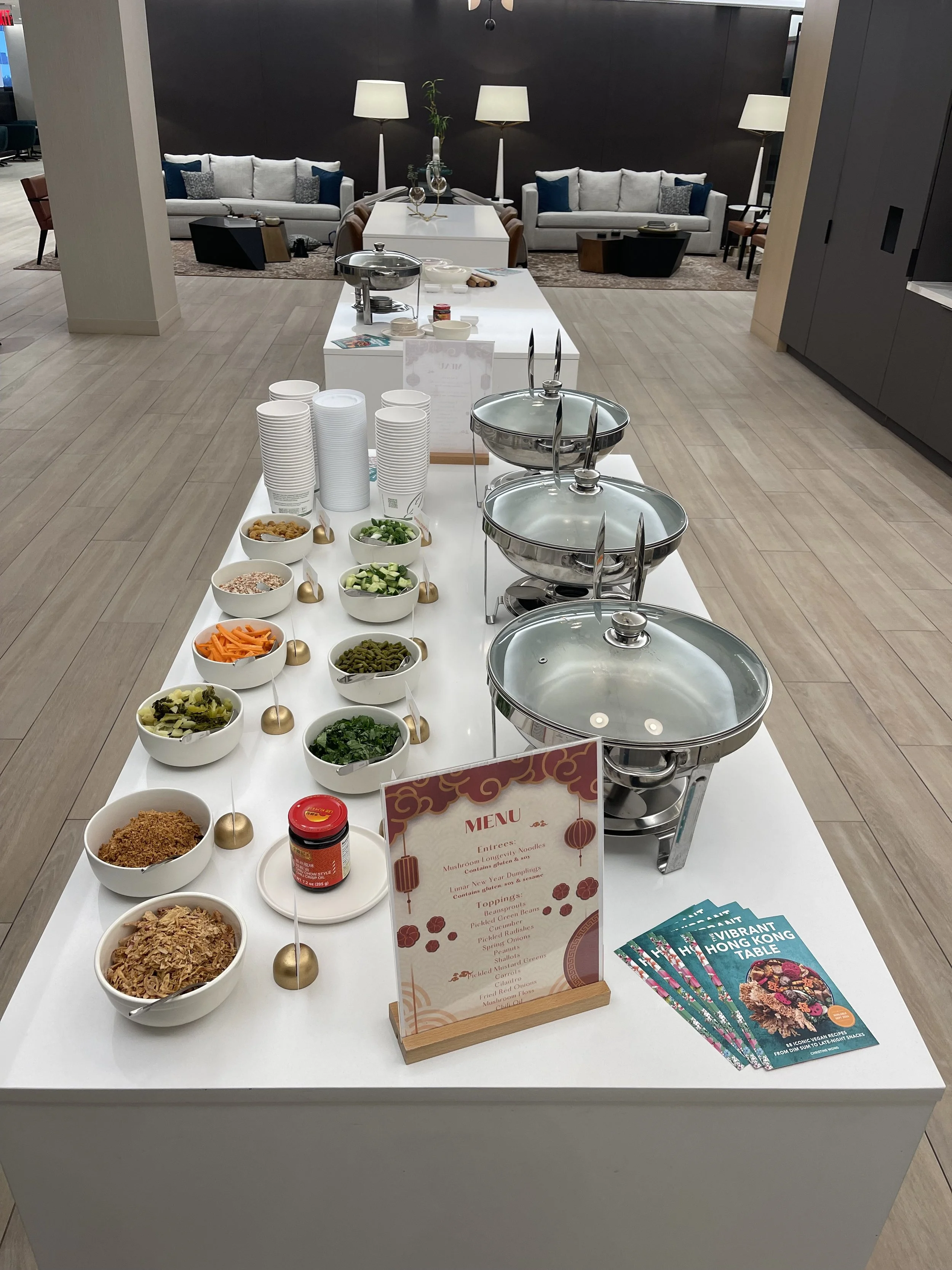 Buffet table with bowls of noodles, vegetables, and condiments, with a menu and flyers nearby, set in a lounge area with sofas and lamps in the background.