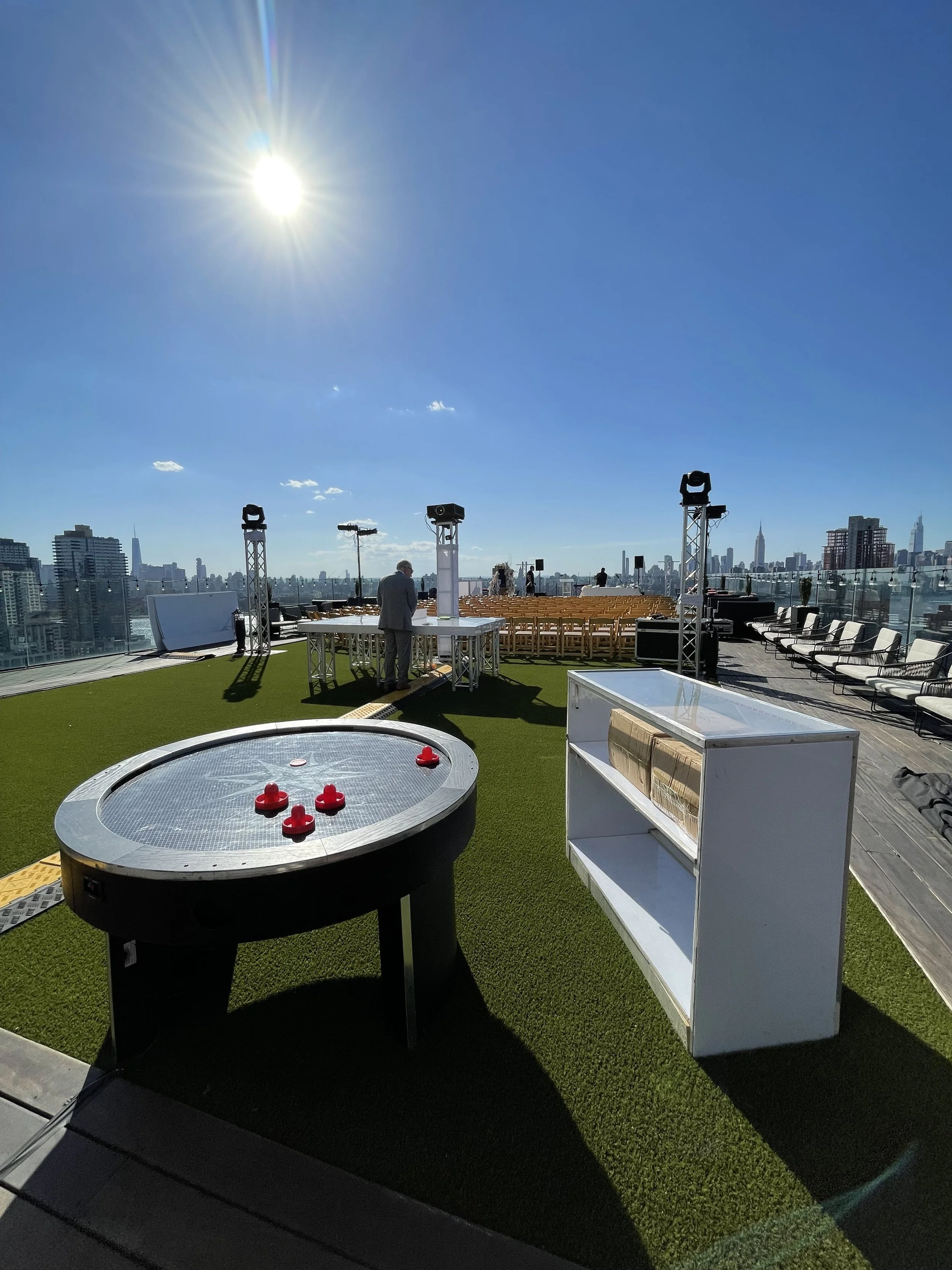 Rooftop event space with city skyline view, artificial grass flooring, seating, audio equipment, and a small fire pit, under a bright sunny sky.