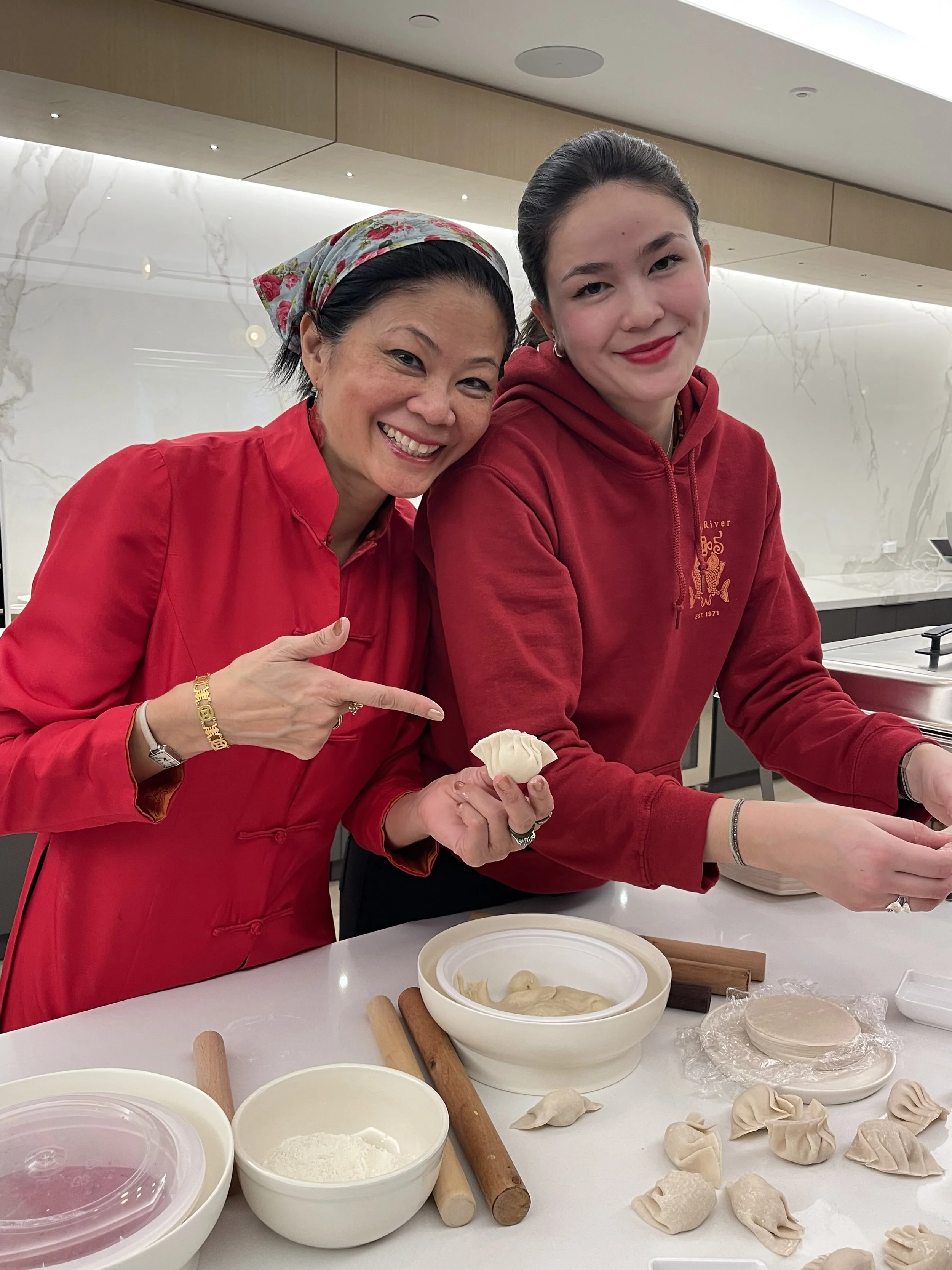 Two women, one older and one younger, making dumplings in a modern kitchen. They are smiling and pointing at the dumplings. The table has various bowls, rolling pins, and dumpling wrappers, with the women wearing red clothing.