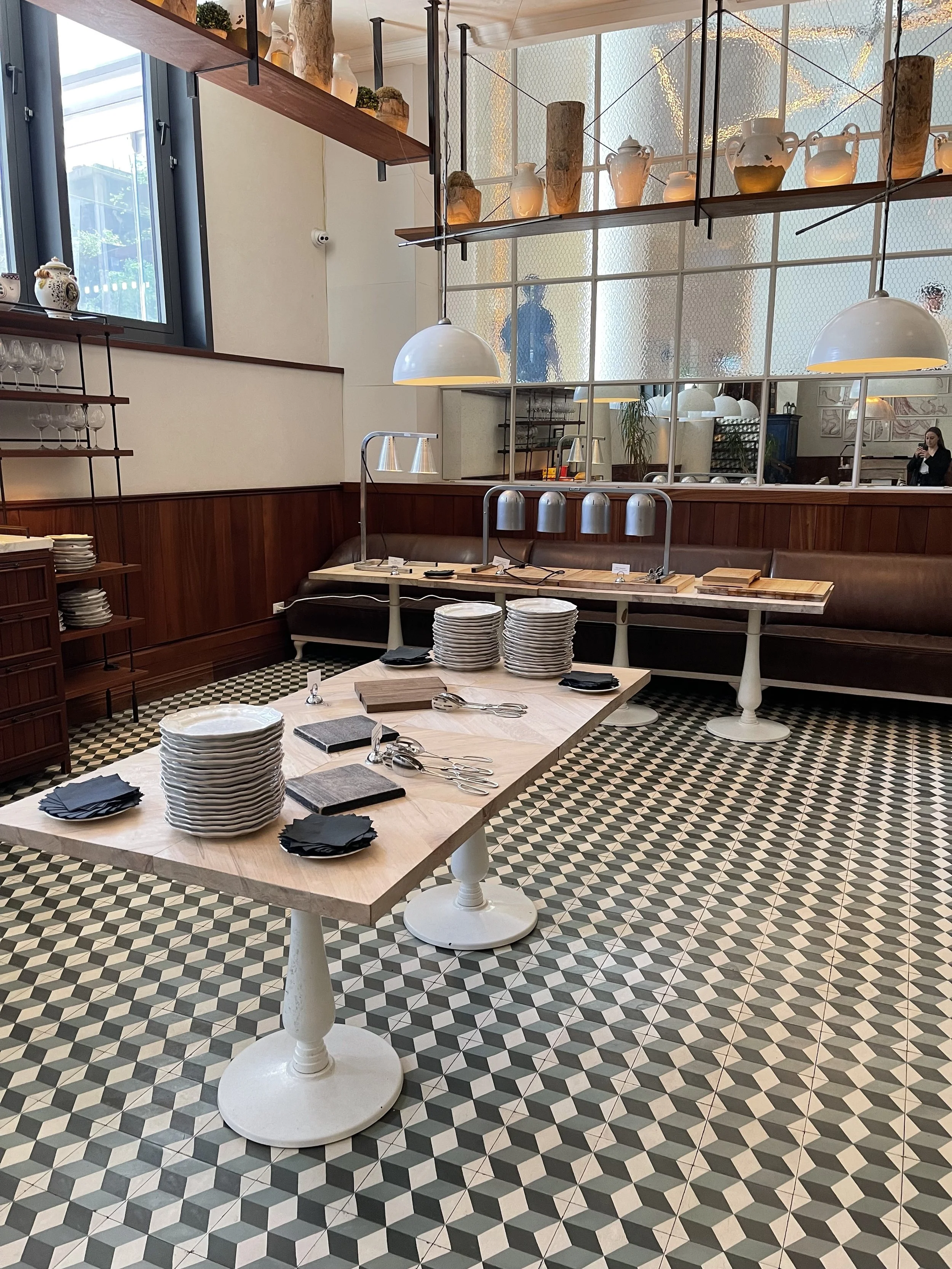 Interior of a modern restaurant or cafe with a patterned black, white, and gray tile floor, a marble table with stacks of plates and black napkins, a wooden bench against the wall, and shelves with decorative vases and pottery. Large windows let in n