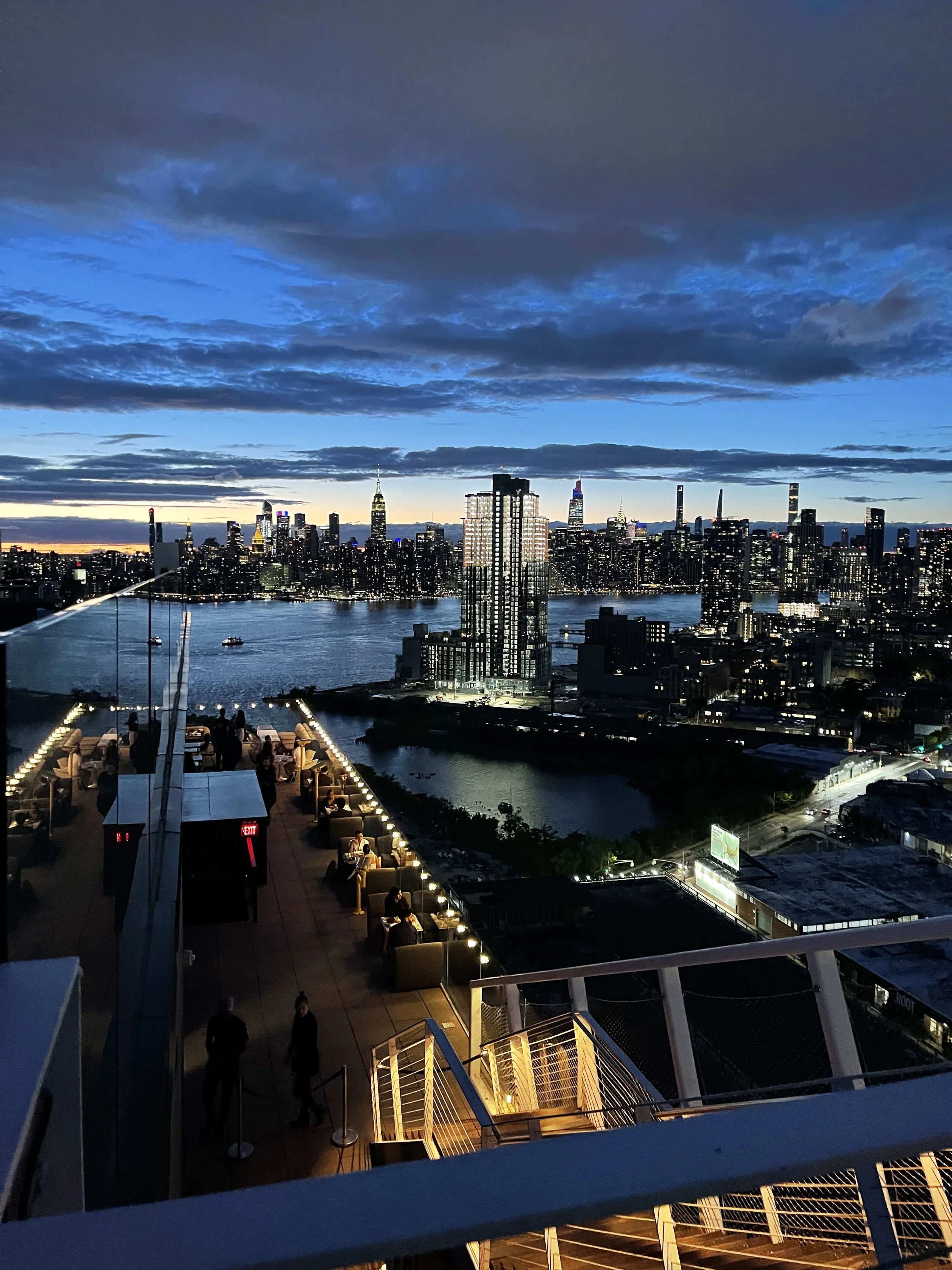 A city skyline at dusk with tall buildings, a river, and a partially cloudy sky. There is a rooftop terrace with people, tables, and string lights.