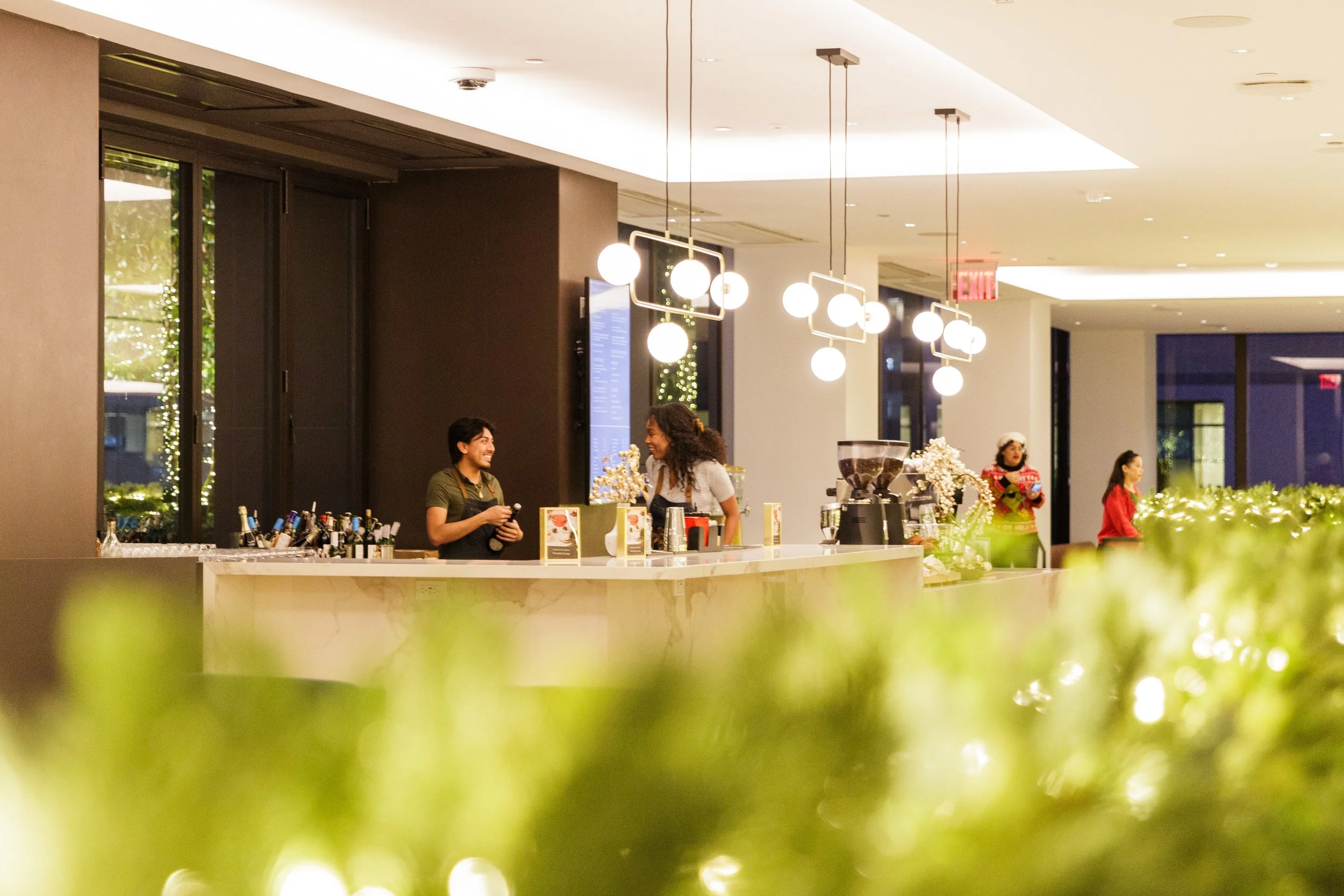 Two women behind a bar counter, smiling and talking, in a modern, well-lit indoor space, possibly a hotel lobby or restaurant, with decorative lighting and people in the background.