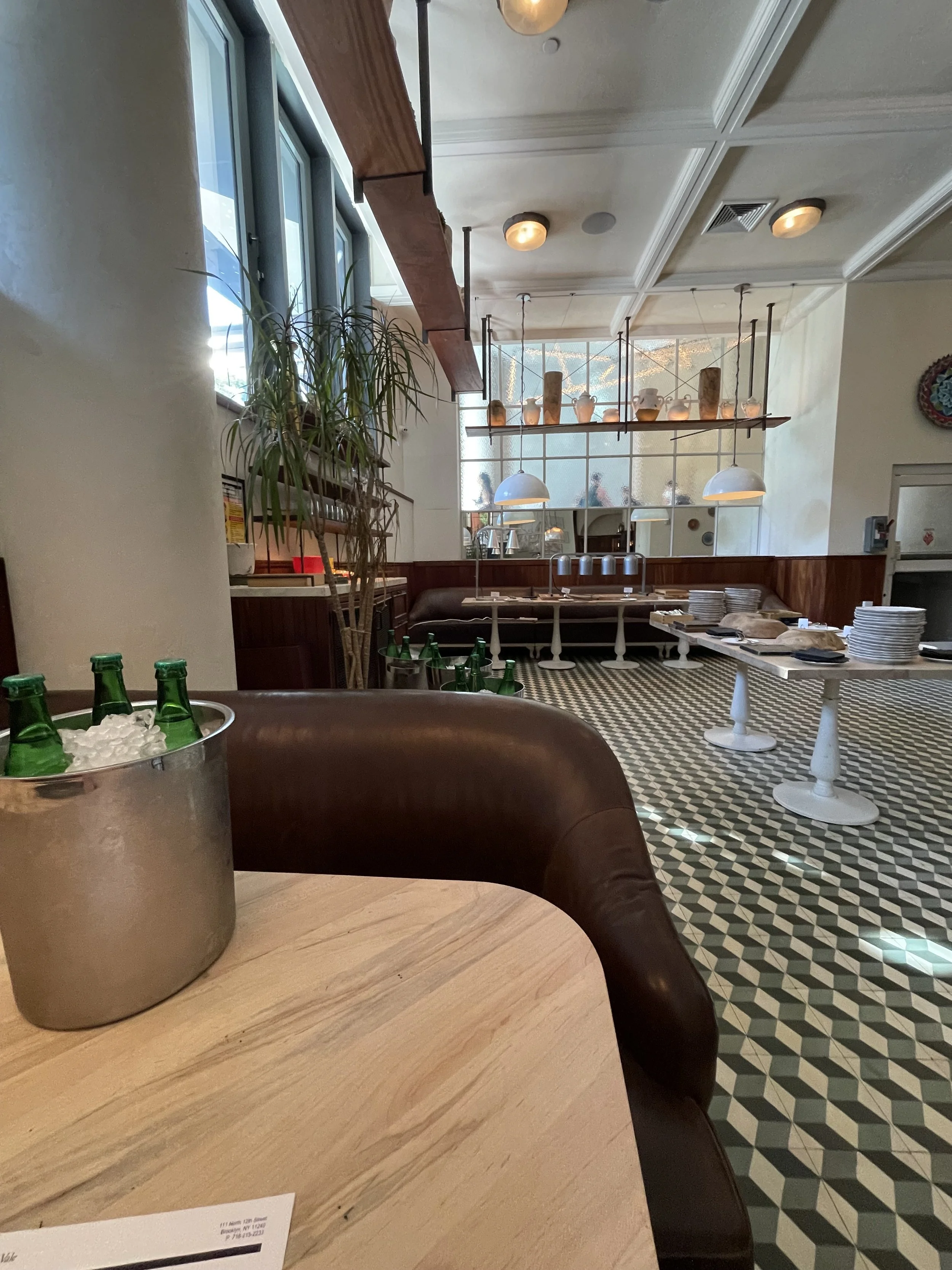 Interior of a restaurant or cafe with a wooden table in the foreground and a chic decor. A potted plant is on the table, and there are stacked plates and bottles. In the background, there are hanging pendant lights, a patterned black and white floor,