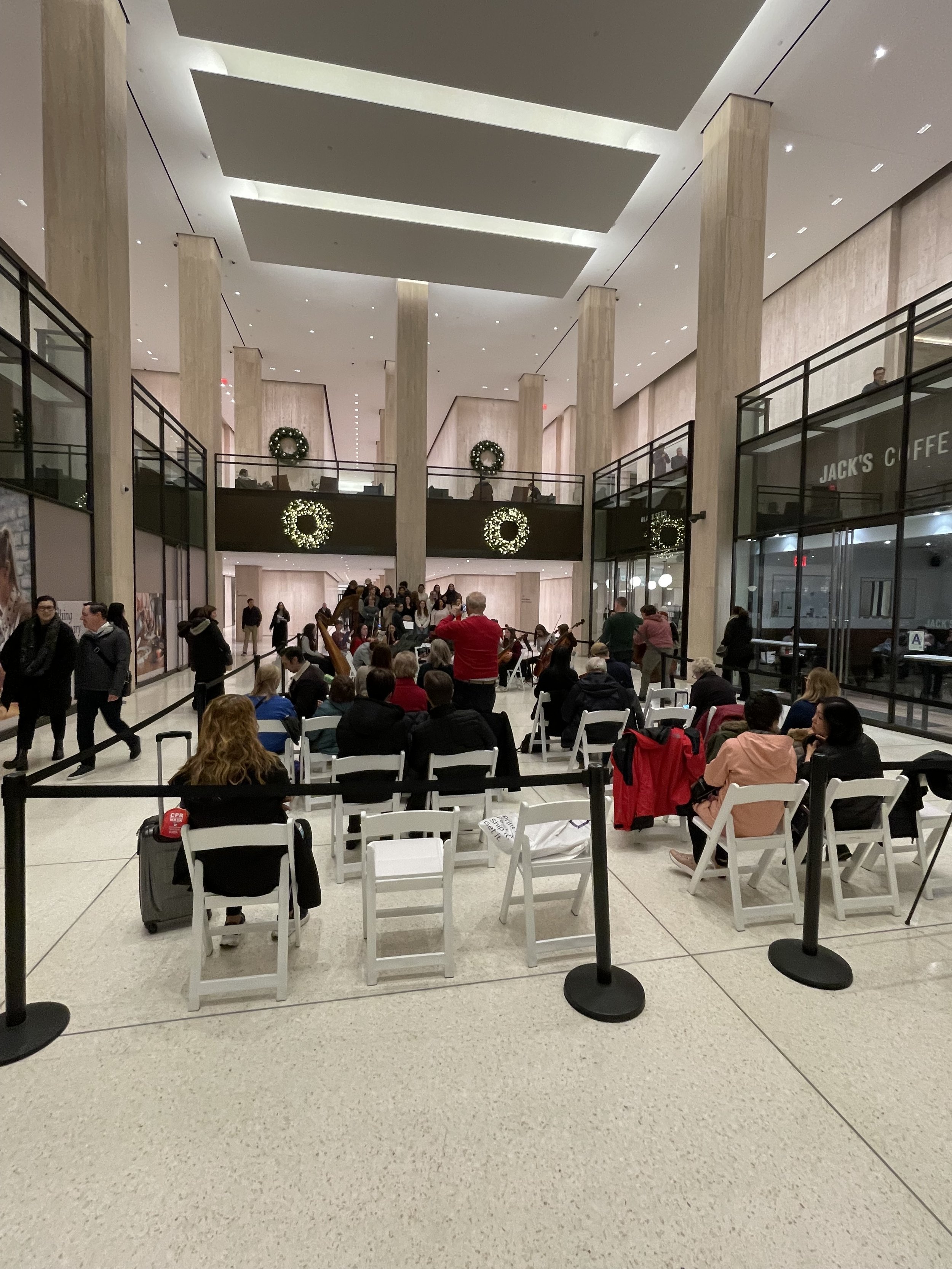 People sitting and waiting in a spacious, modern indoor space, likely an airport terminal, with a musicians performing in the background, decorated with holiday wreaths.