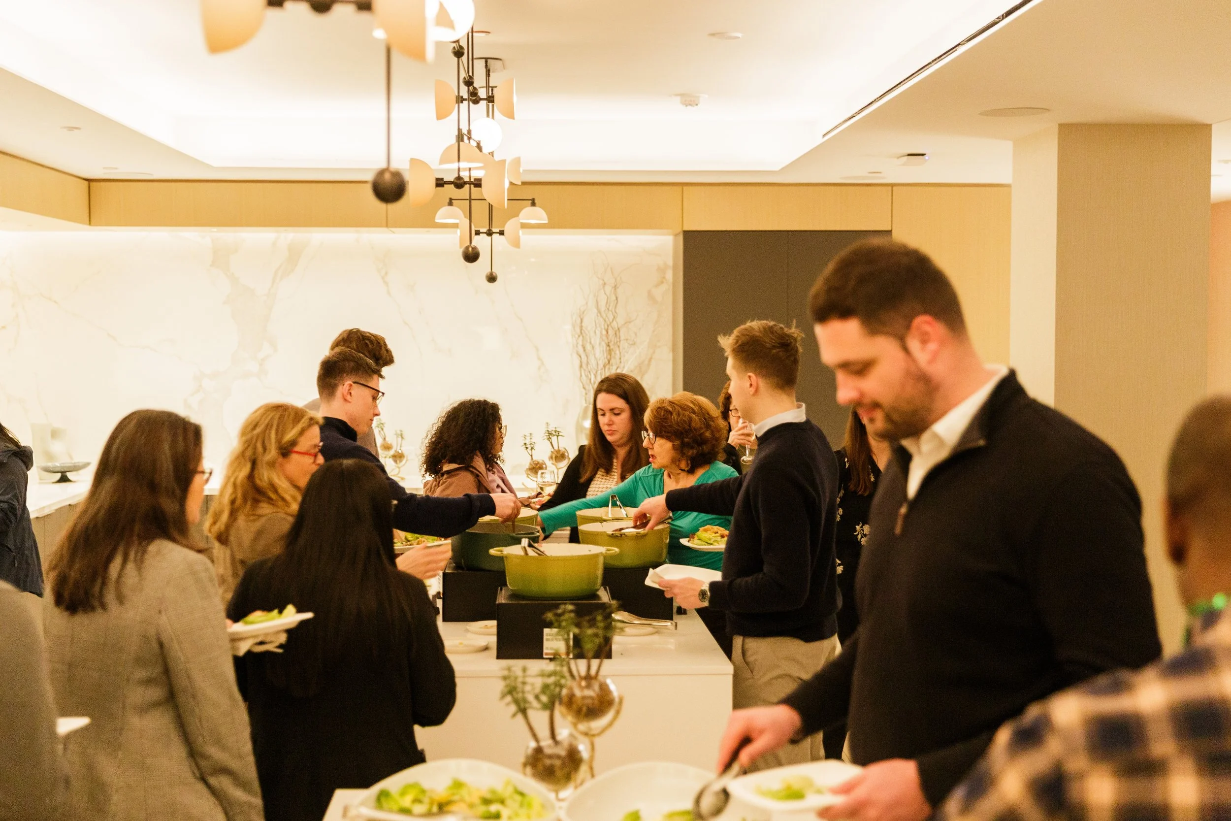 A group of people at a buffet table in an upscale restaurant or hotel, selecting food from various dishes, with modern decor and warm lighting.
