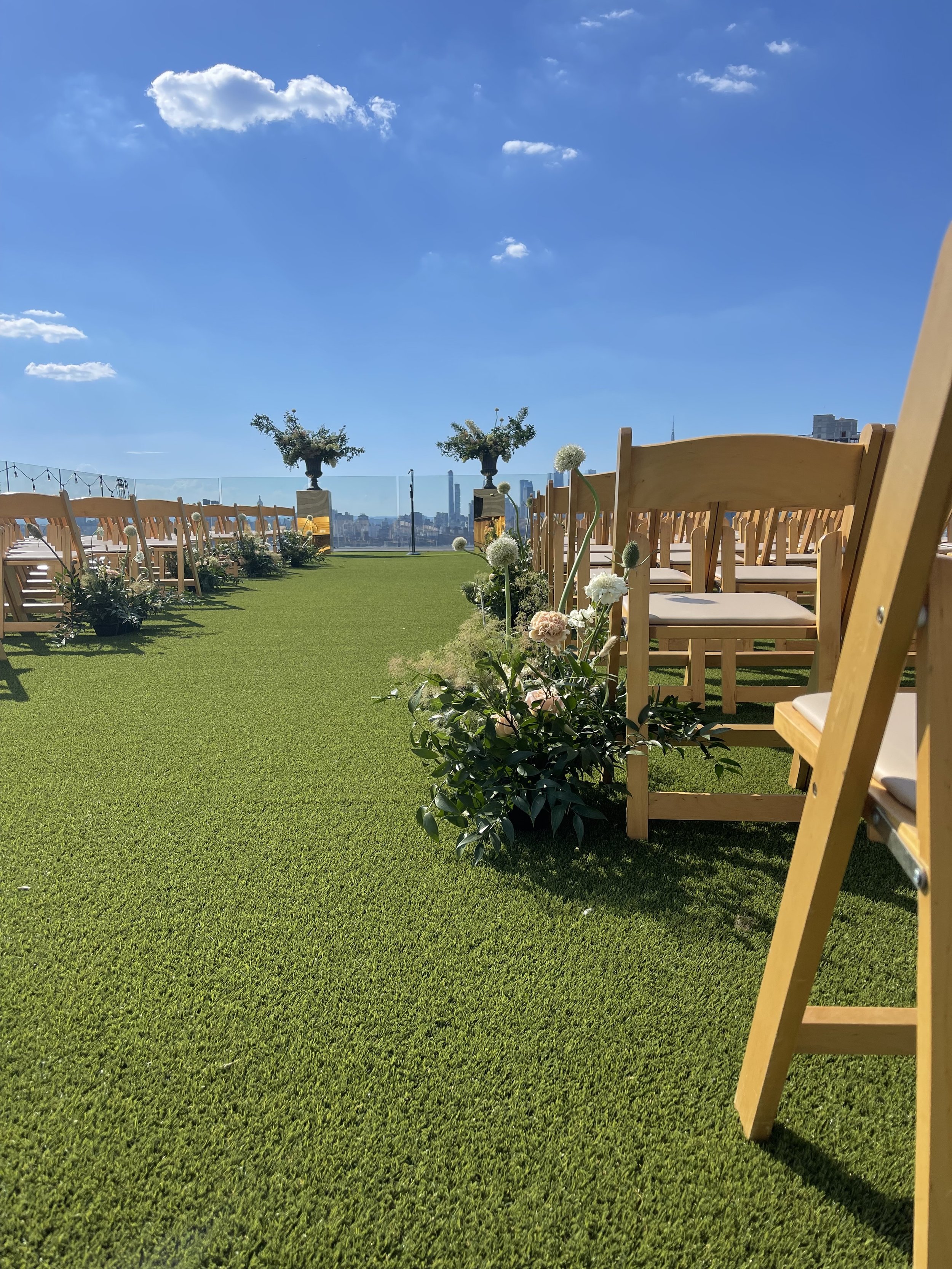 Outdoor event space with wooden chairs arranged on a green lawn, adorned with flowers and decorative trees, under a blue sky with white clouds, with a city skyline in the background.