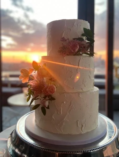 A two-tiered white wedding cake with floral decorations, set against a sunset background through window, displayed on a silver cake stand.
