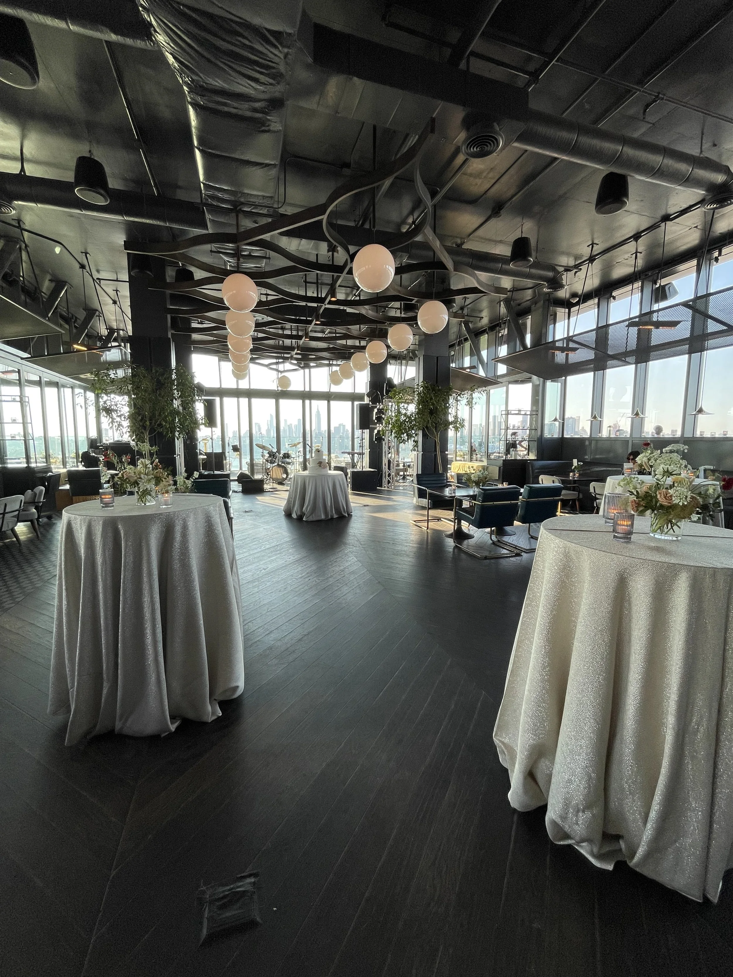 Indoor event space with decorated tables, large windows showing city skyline, modern ceiling design with hanging globe lights, musical instruments on stage, potted plants, and a mix of lounge chairs and tables for seating.