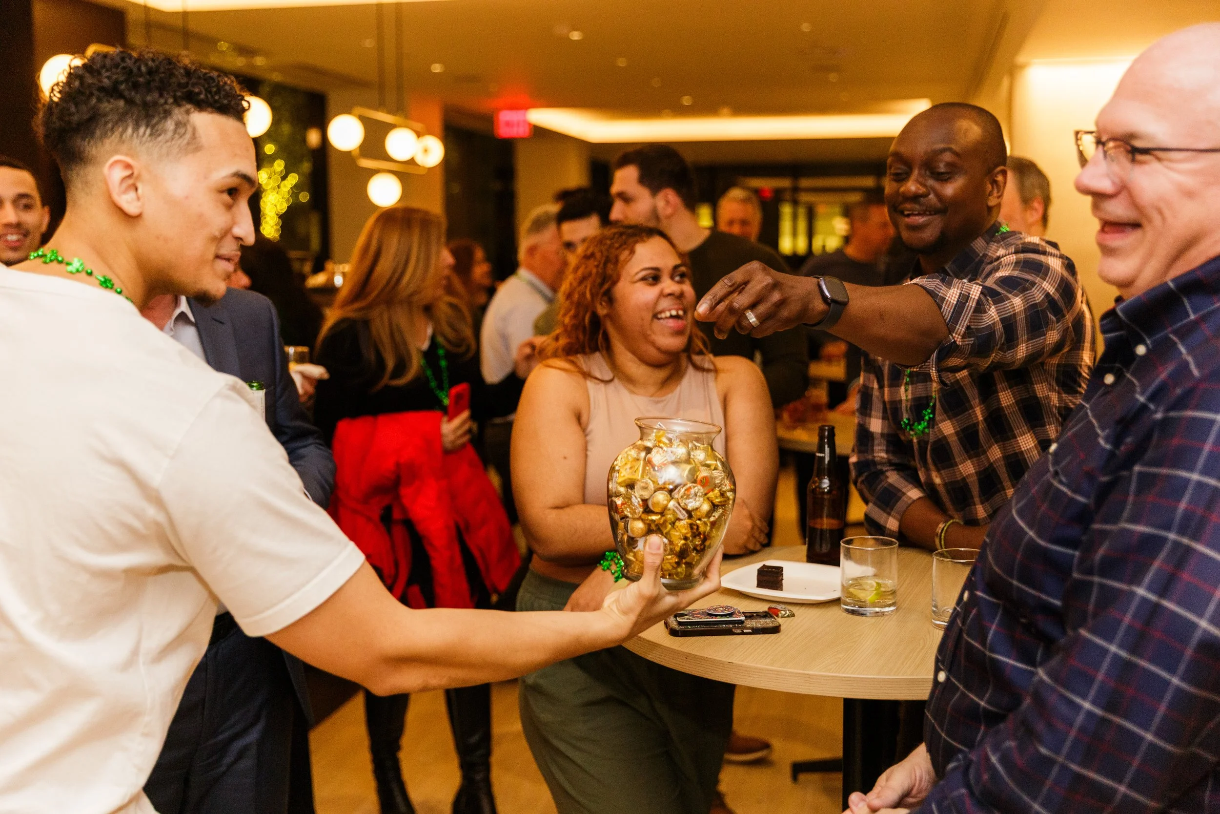 A group of people socializing at a party, with some guests wearing green bead necklaces, and one woman holding a vase filled with gold-wrapped candies.