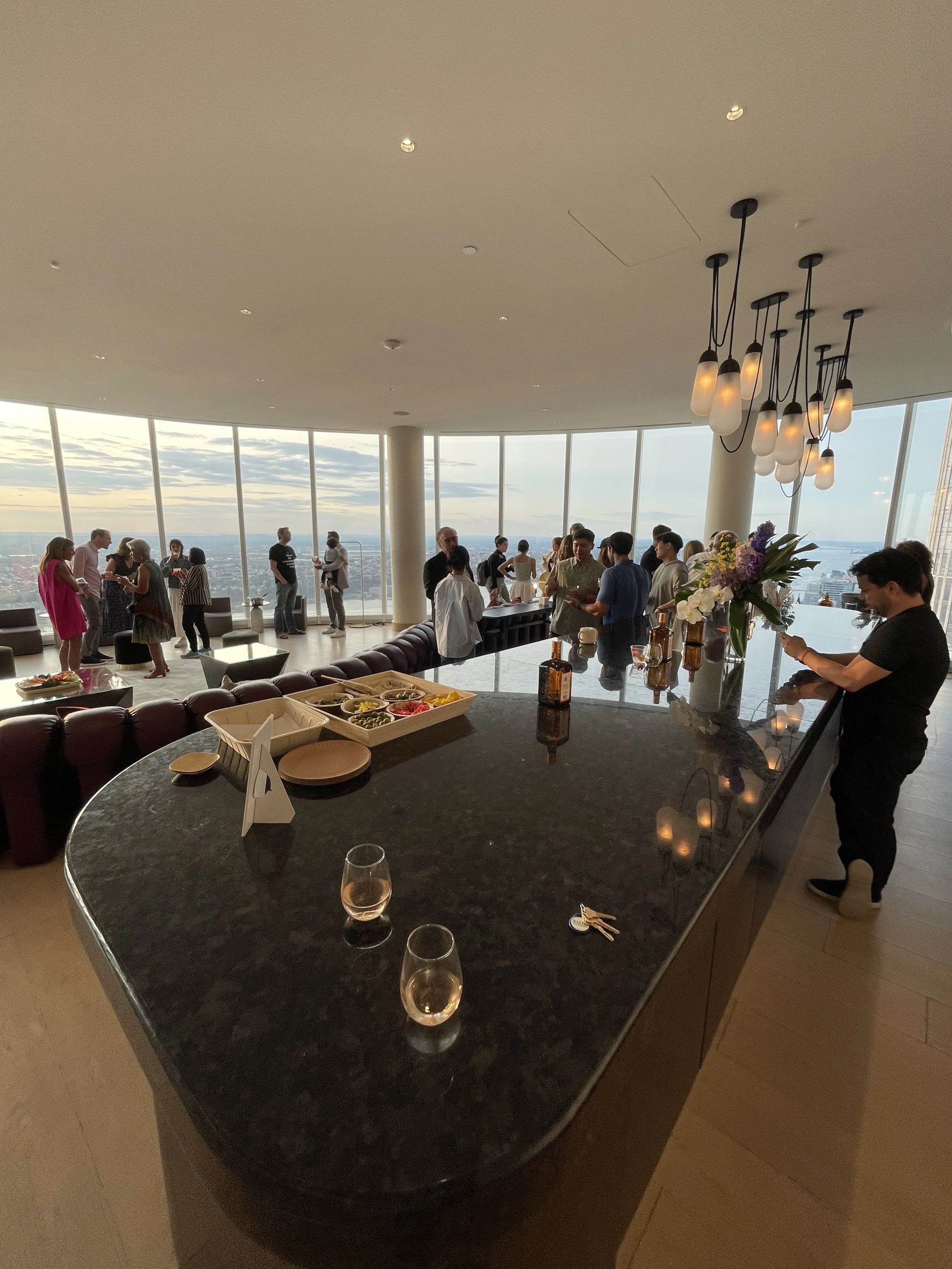 Interior of a high-rise building with floor-to-ceiling windows showing a cityscape and water. People are gathered, chatting and mingling. A large black marble countertop with a glass of rosé wine, some bottles, and a flower arrangement is in the fore