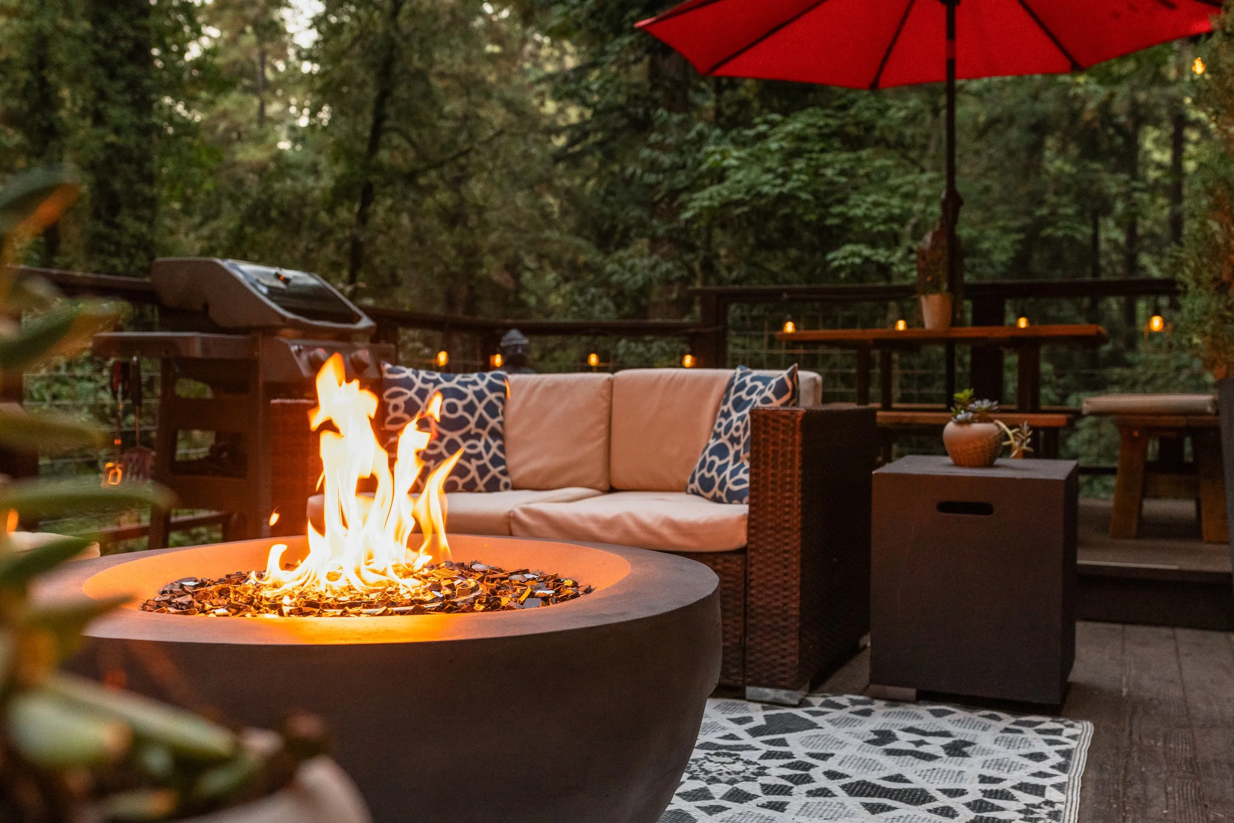 An outdoor patio at dusk with a fire pit, a beige sofa with patterned blue and white pillows, a side table with a potted plant, and a red umbrella among lush green trees.