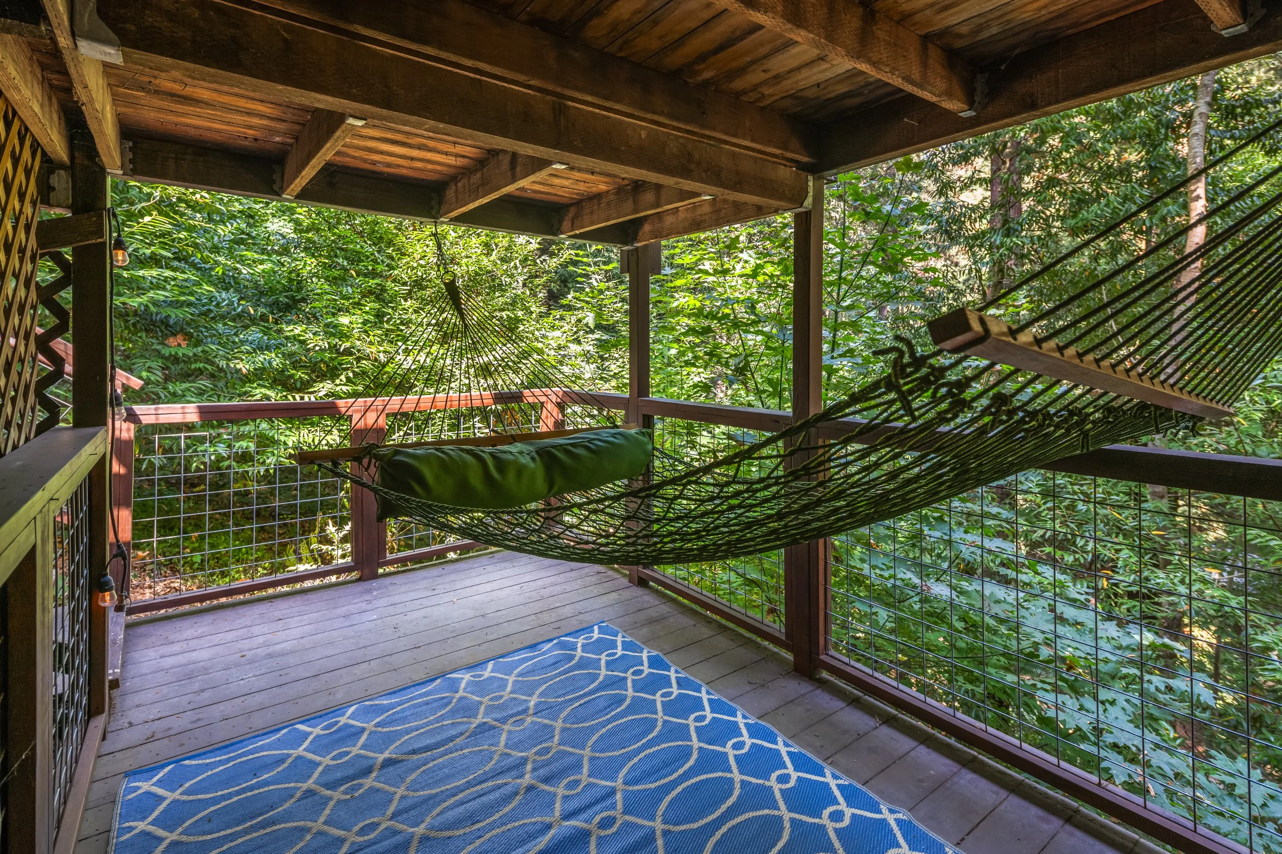 A wooden deck with a blue patterned outdoor rug, a green hammock, and surrounded by green trees and foliage, with parts of the wooden railing visible.