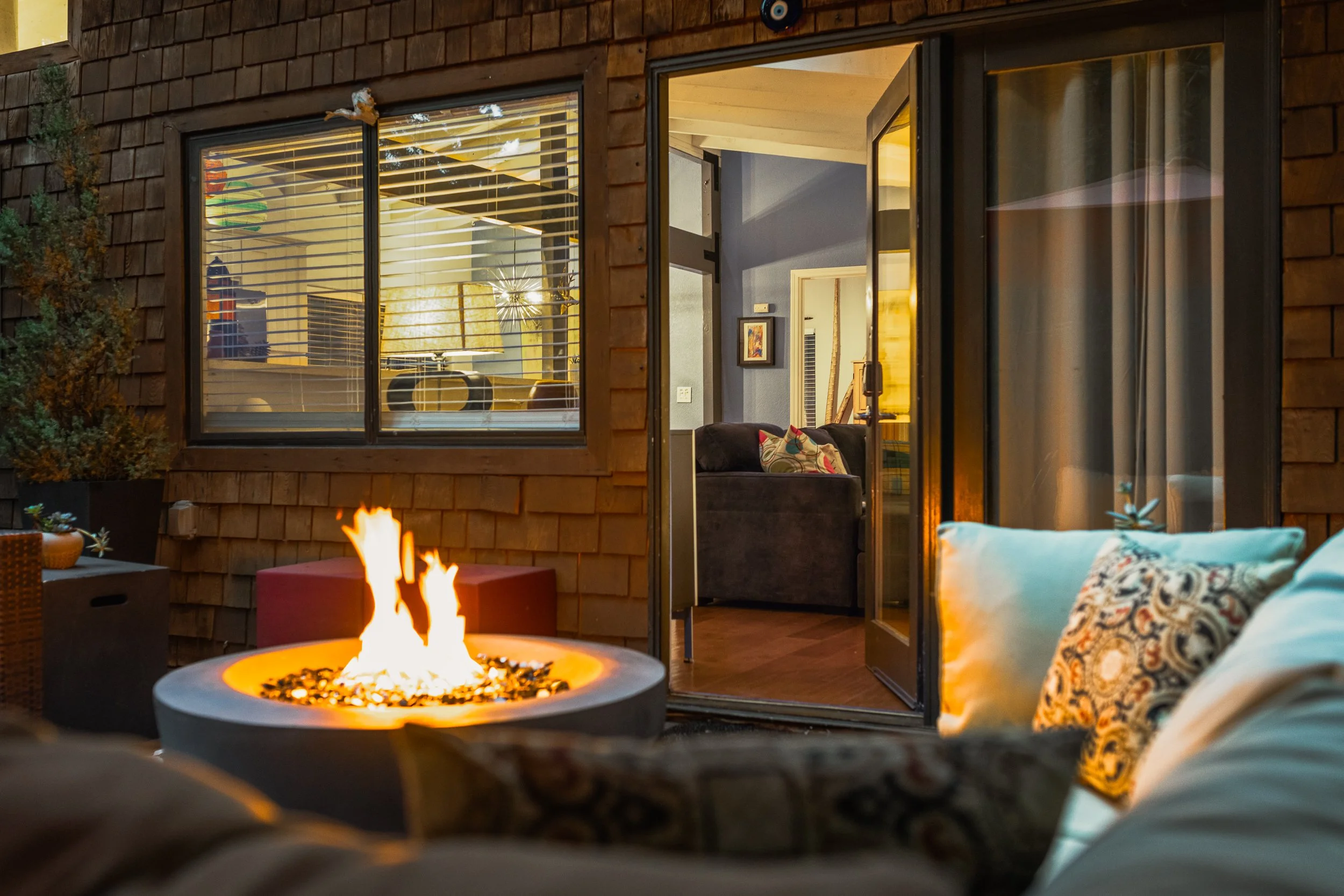 A cozy outdoor patio with a fire pit, a sofa with patterned cushions, and a house with large windows showing a living room inside. The patio has plants and outdoor furniture, creating a warm evening ambiance.